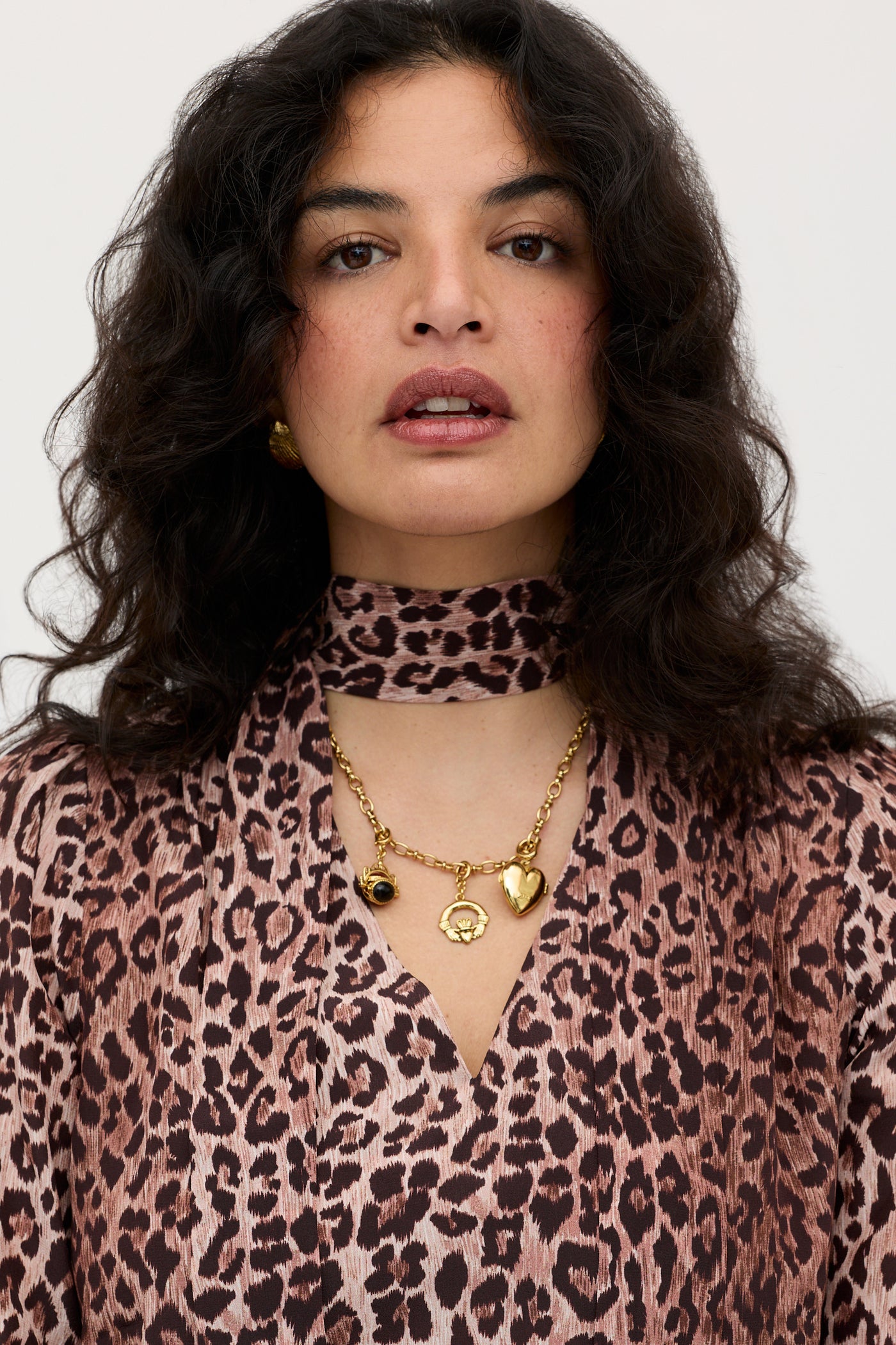 A woman with wavy dark hair models the Moss - Leopard Brown top, a vintage-inspired silk piece in brown and pink leopard print with a matching scarf. She pairs it with gold earrings and a charm necklace, facing the camera against a plain background.
