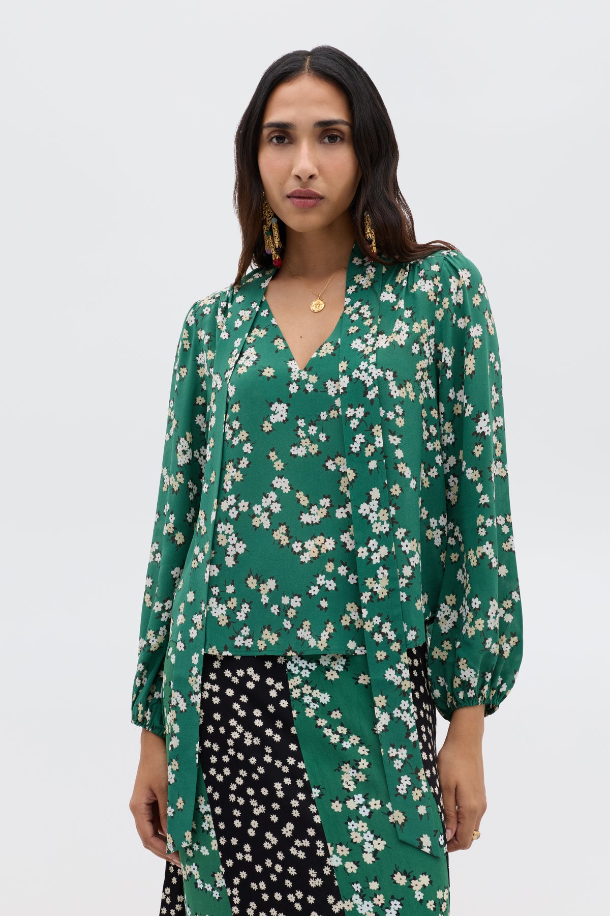 A woman wears the Moss - City Floral Olive blouse, a vintage-inspired green top with white floral print and v-neckline, paired with a black patterned skirt. She has long dark hair, gold jewelry, and a plain white background.