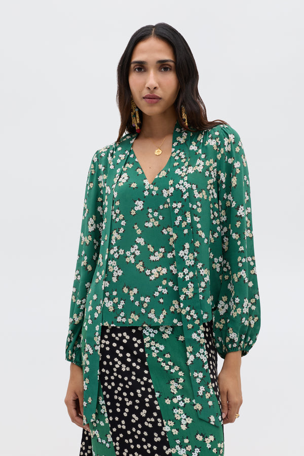 A woman wears the Moss - City Floral Olive blouse, a vintage-inspired green top with white floral print and v-neckline, paired with a black patterned skirt. She has long dark hair, gold jewelry, and a plain white background.