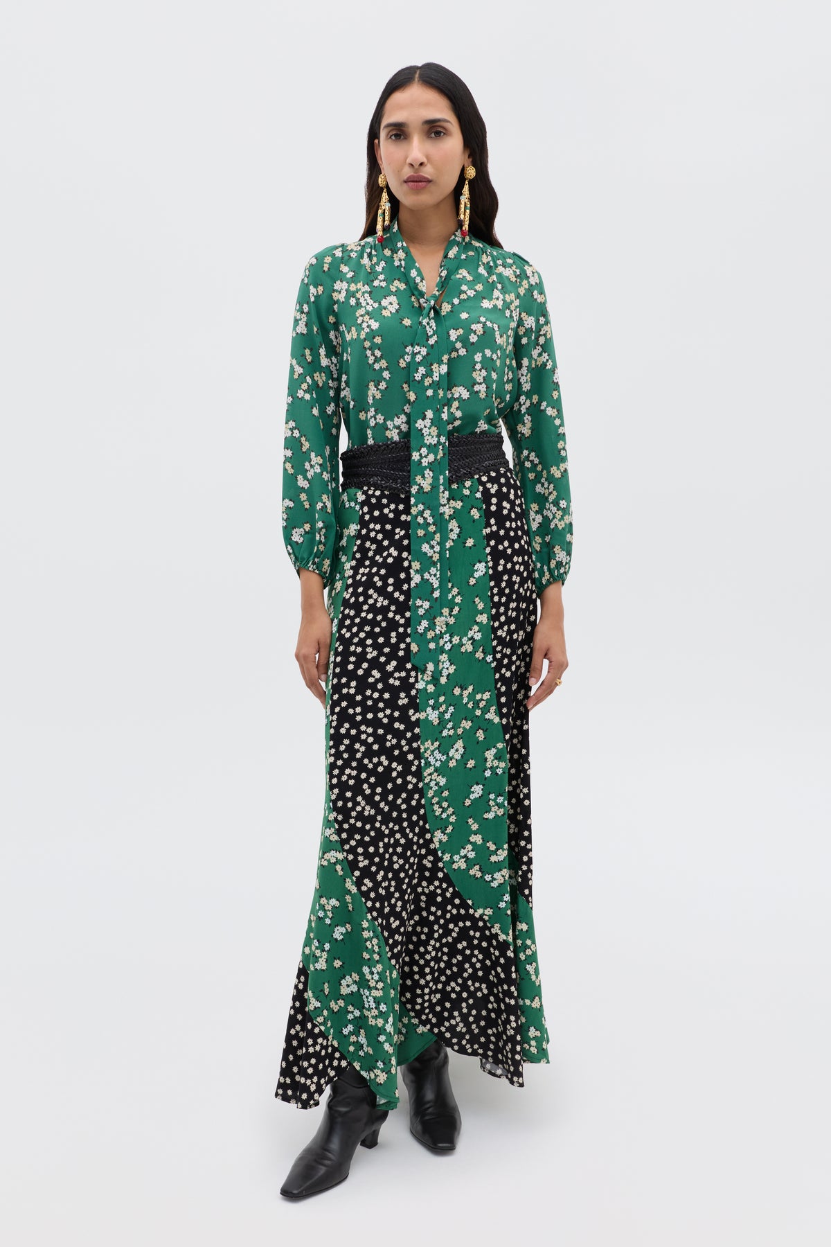 A woman faces forward wearing the Moss - City Floral Olive green and black floral silk Crepe de Chine long-sleeve maxi dress with a black belt, black ankle boots, and gold earrings against a plain white background.