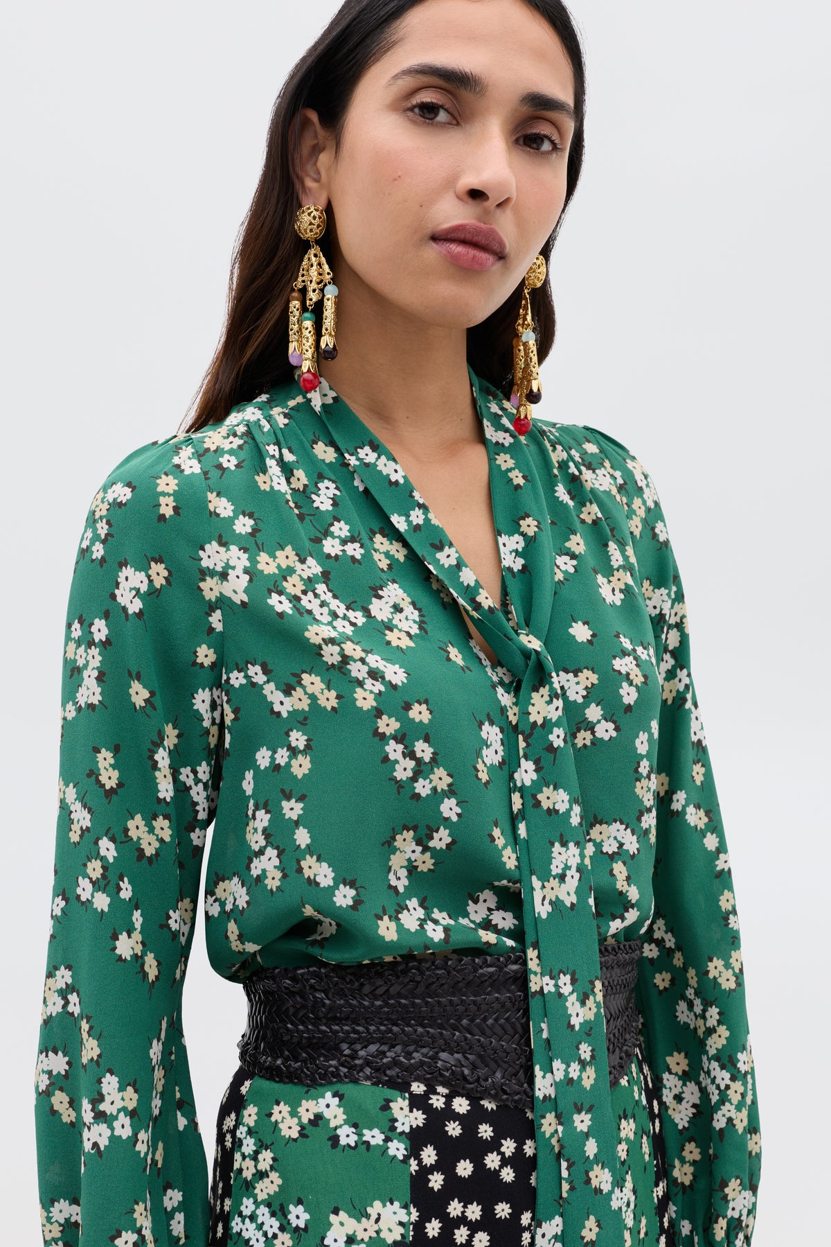 A woman wears the Moss - City Floral Olive blouse, a green floral silk Crepe de Chine with a scarf neckline, paired with a black woven belt and ornate gold earrings, standing against a plain light background.