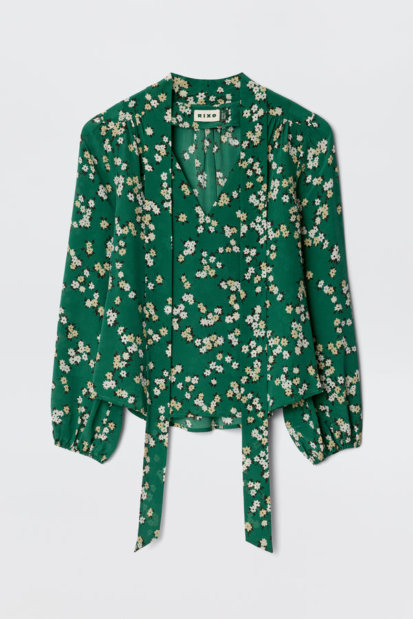 The Moss - City Floral Olive is a vintage-inspired green silk blouse with white and yellow florals, a v-neck, gathered shoulders, and two long front ties.