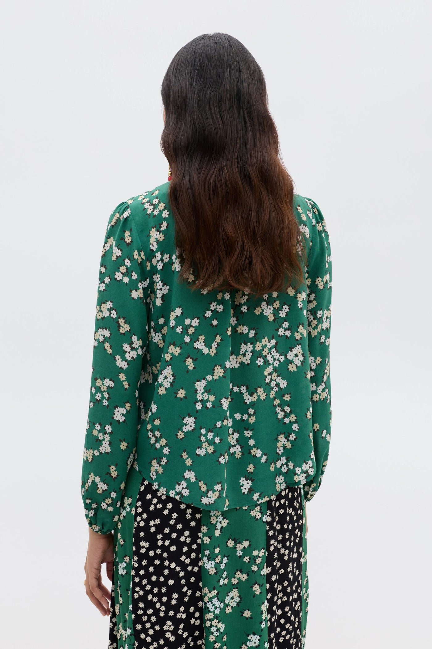 Facing away, a person with long wavy hair wears the Moss - City Floral Olive vintage-inspired green silk Crepe de Chine blouse with a scarf neckline, paired with a black skirt with white flowers against a plain light background.