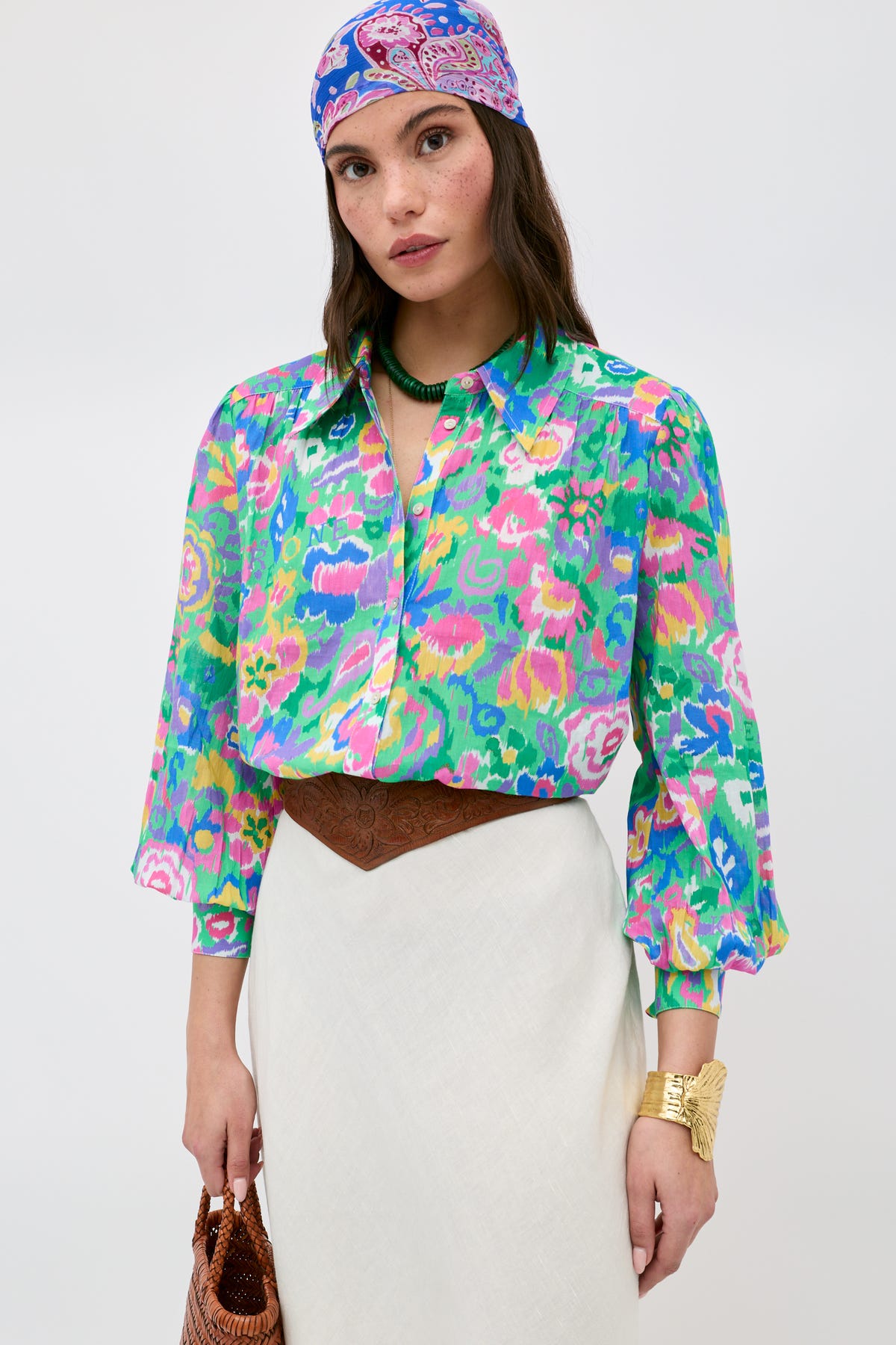 A woman stands against a plain background, wearing the Blake - Ikat Bloom Green floral blouse paired with a white skirt, patterned headscarf, brown belt, gold bracelet, green necklace, and holding a brown bag.
