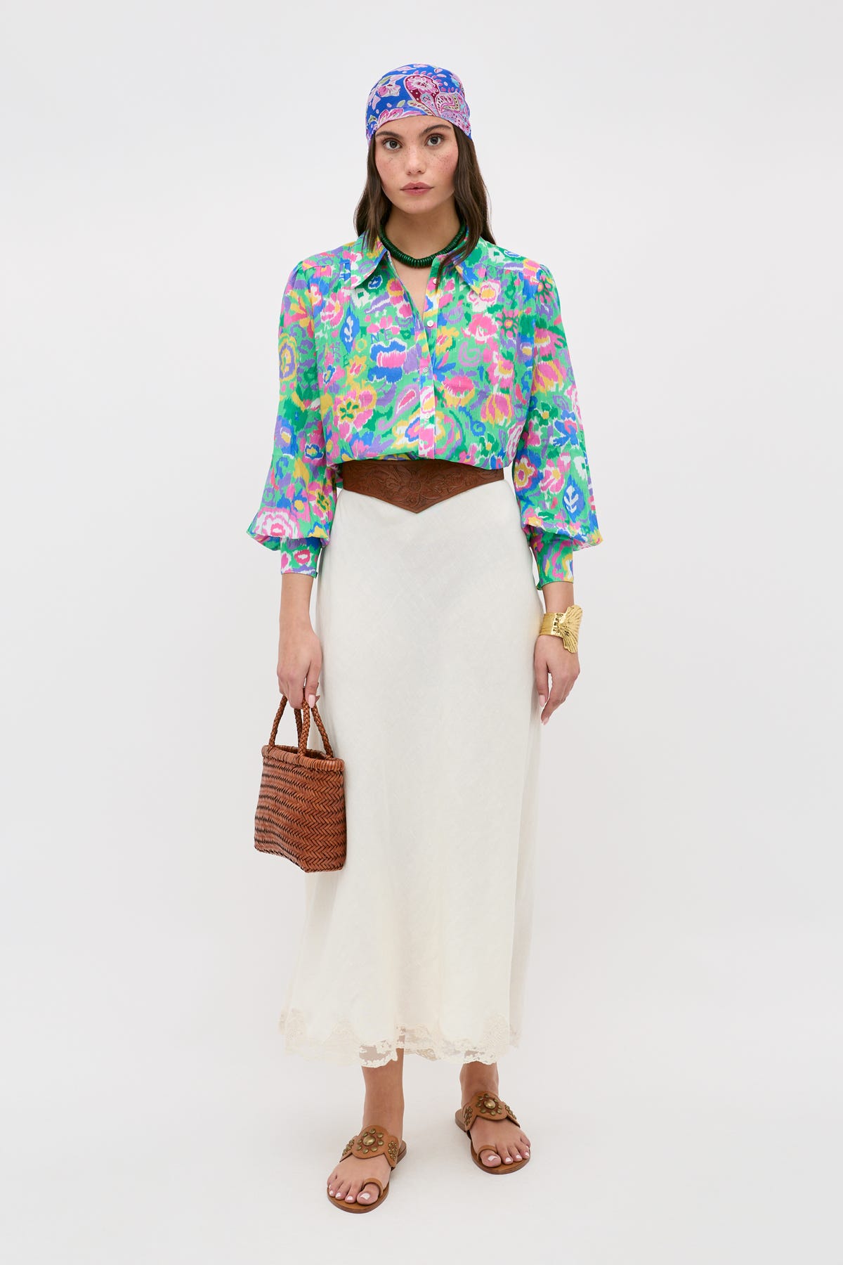 A woman wears the Blake - Ikat Bloom Green top, paired with a white skirt, brown belt, sandals, and a coordinating headscarf. She carries a woven brown handbag and poses with a neutral expression against a plain background.