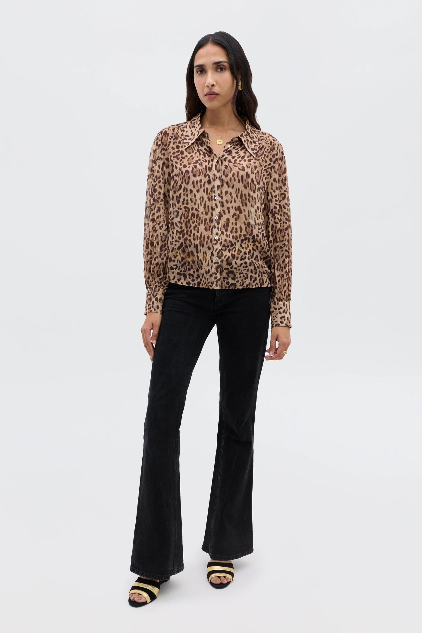 A woman with long dark hair and a neutral expression stands against a white background, wearing the Blake - Mila Leopard Brown top, black flared pants, and black open-toe heeled sandals.