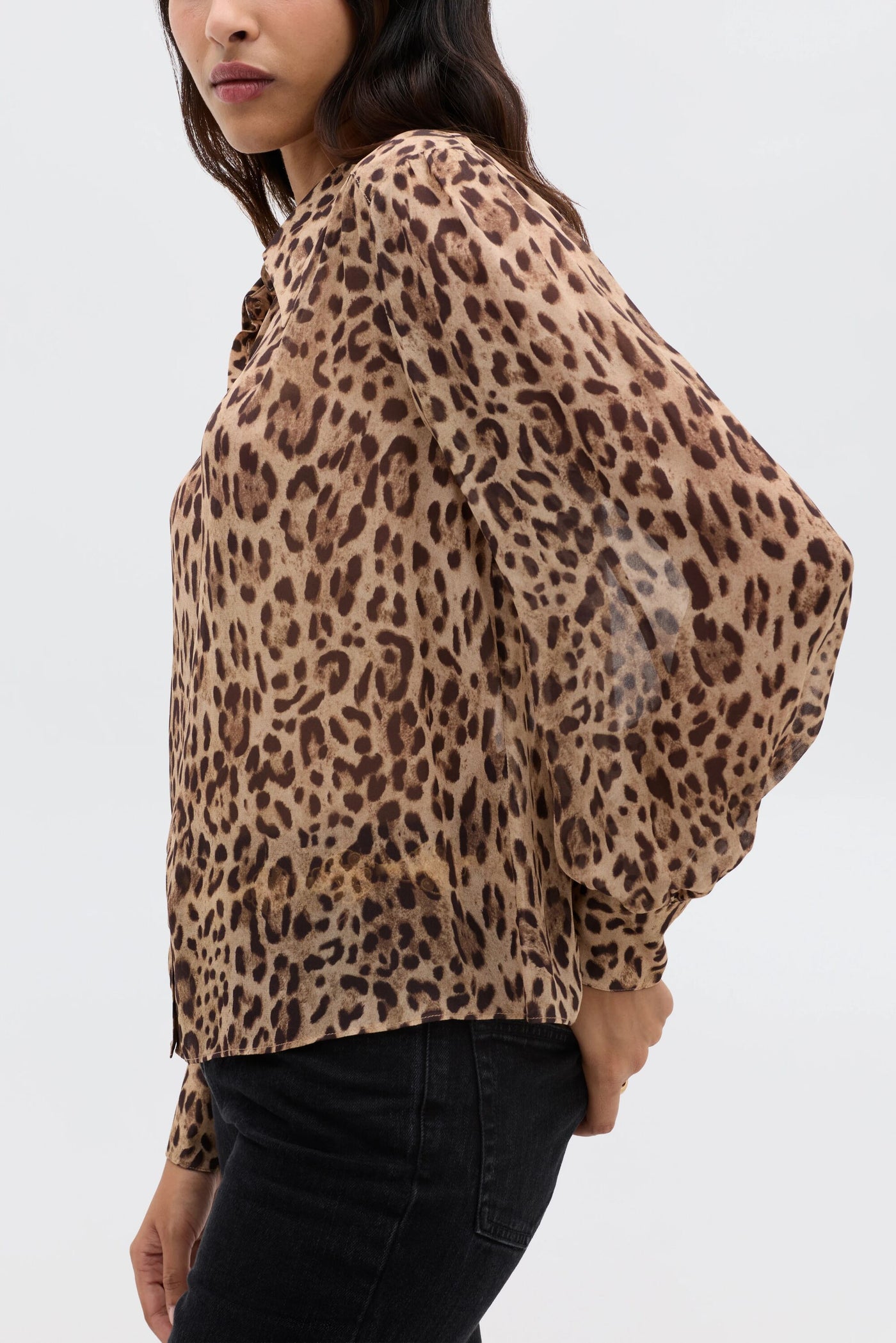 A woman stands sideways against a plain light background, wearing the Blake - Mila Leopard Brown blouse with black jeans and one hand behind her back.