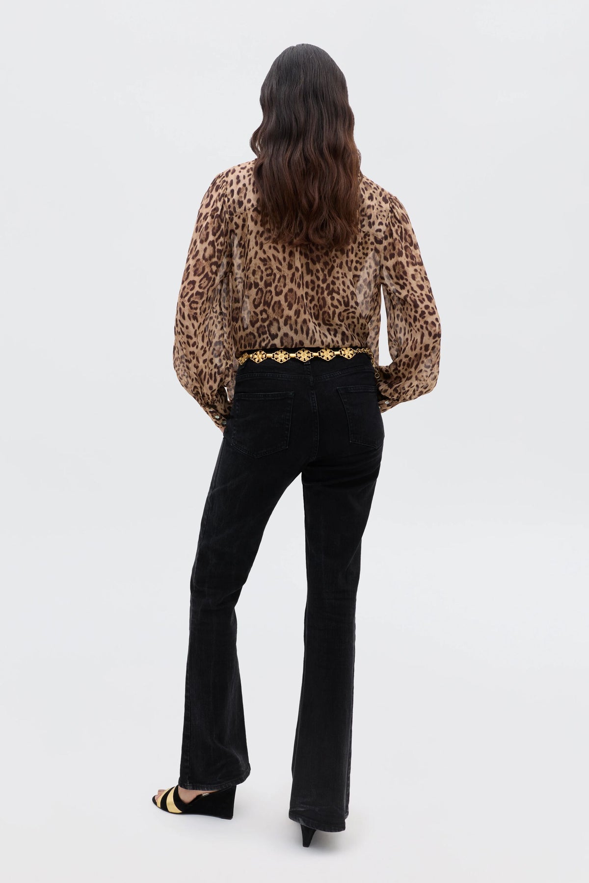 A person with long wavy hair faces away, wearing the Blake - Mila Leopard Brown vintage-inspired top, black pants, black heels, and a gold chain belt against a plain white background.
