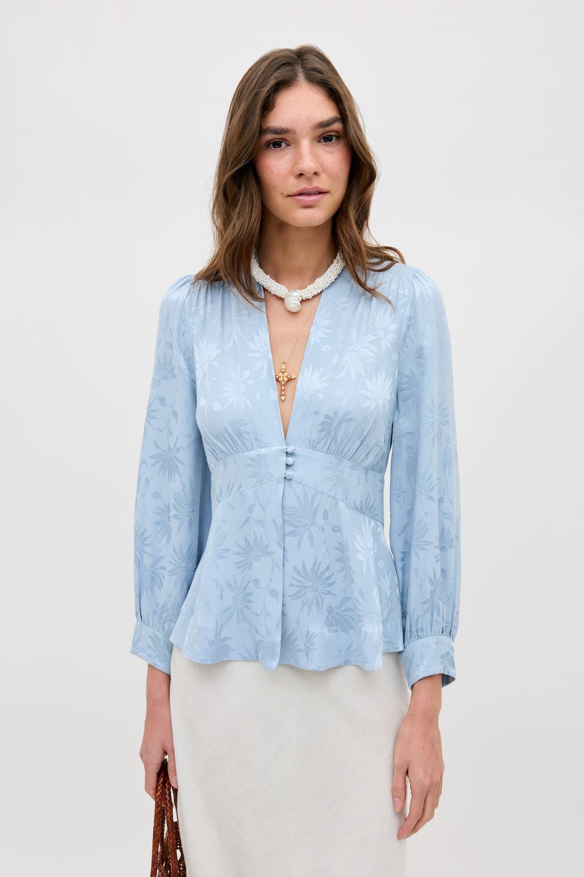 A woman stands against a plain background wearing the Lea - Daisy Jacquard Bluebell, a light blue, long-sleeve blouse with a floral pattern and deep V-neck. She holds a brown woven bag and accessorizes with a white necklace featuring a pendant.