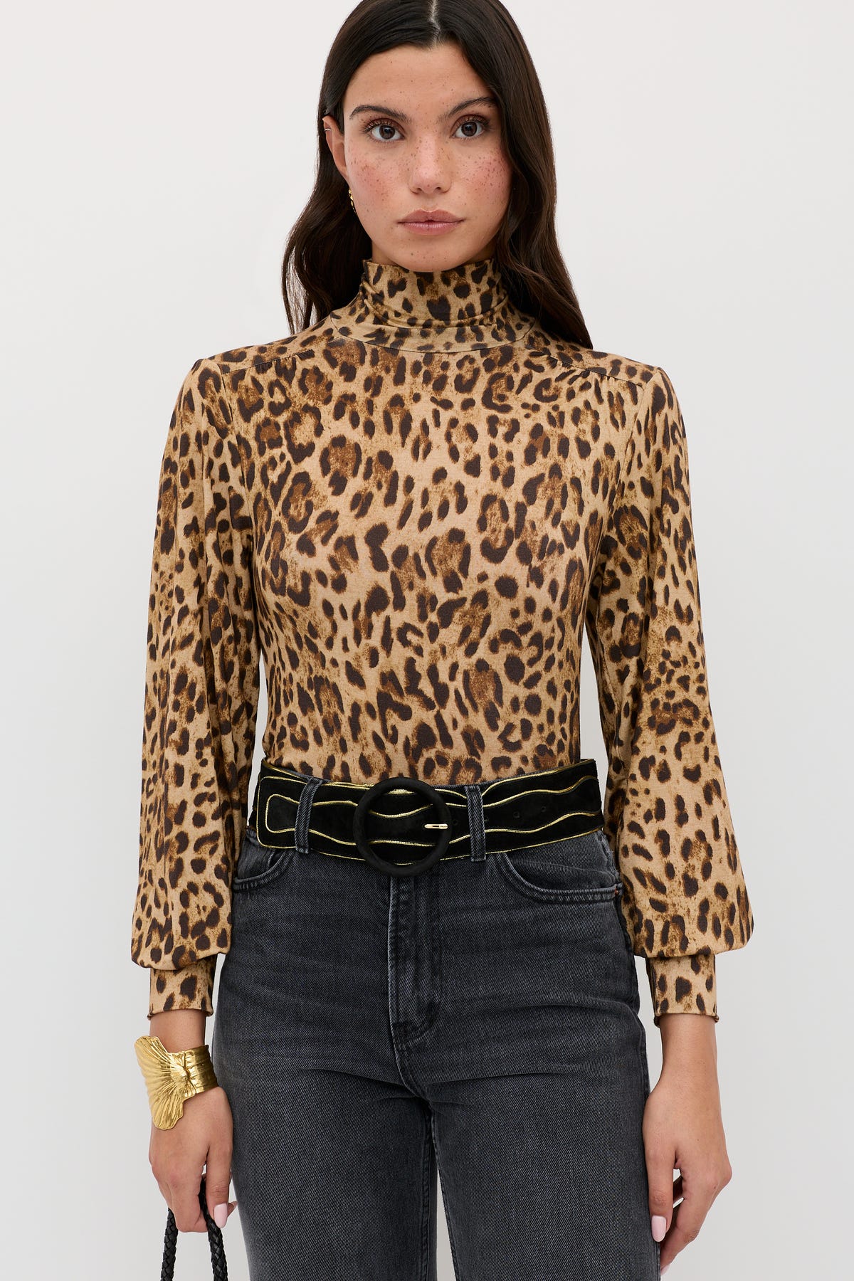 Woman wearing the Suki - Mila Leopard Brown turtleneck layering top, black high-waisted jeans, and a black belt with a gold buckle. She has long, dark hair and holds a small black bag.