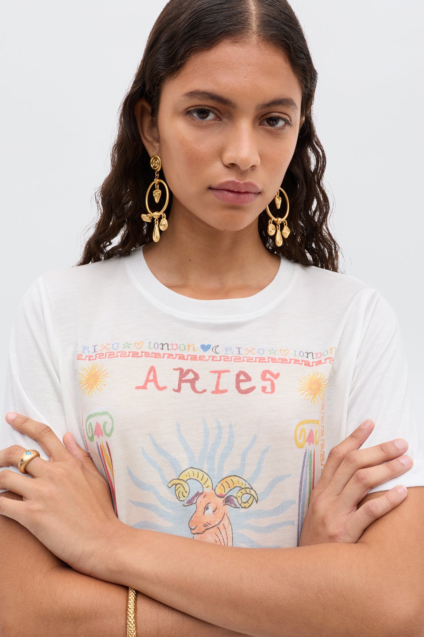 A woman with long wavy hair wears gold earrings and jewelry, plus a white Ria - Aries T-shirt featuring a colorful Aries illustration with a ram graphic and cropped hem. She stands with arms crossed, looking at the camera.