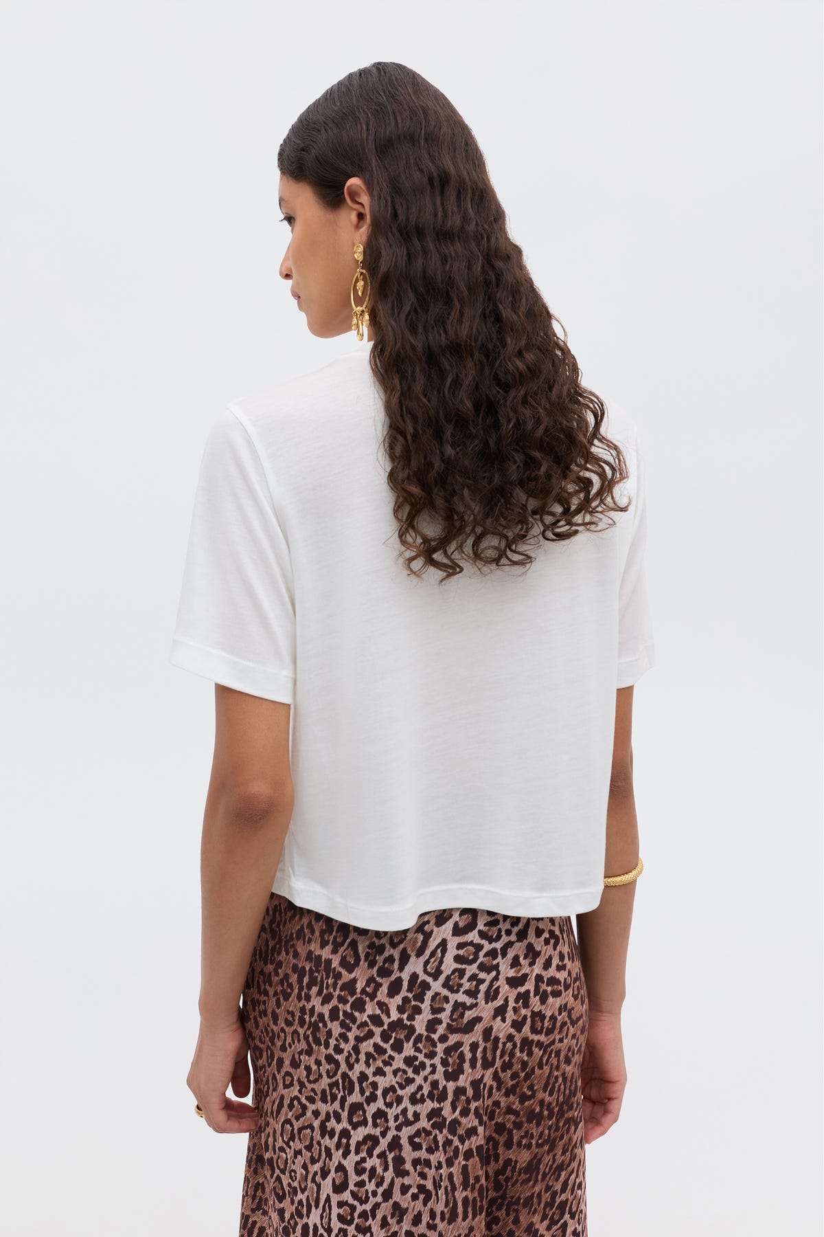 A woman with long, wavy dark hair is seen from behind in a white cropped tee and the brown leopard-print Ria - Taurus skirt by RIXO, accessorized with gold earrings and bracelets against a plain light background.