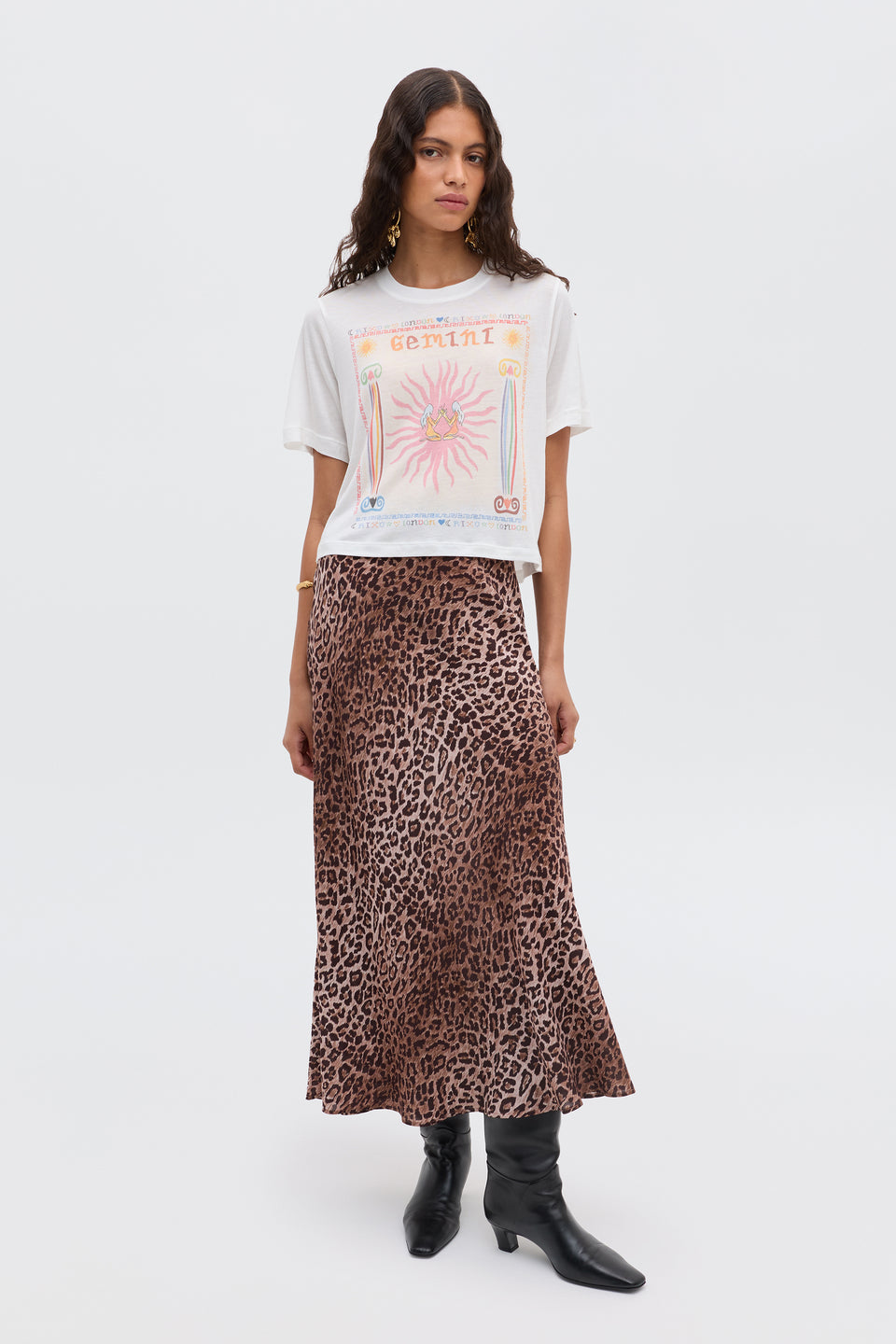 A woman stands against a plain background wearing the Ria - Gemini T-shirt in white, styled with a brown leopard print midi skirt and black pointed-toe boots.