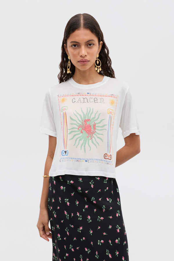 A woman with long, wavy hair and gold hoop earrings wears the white Ria - Cancer T-shirt featuring a colorful Cancer illustration, paired with a black floral-print skirt, against a plain background.