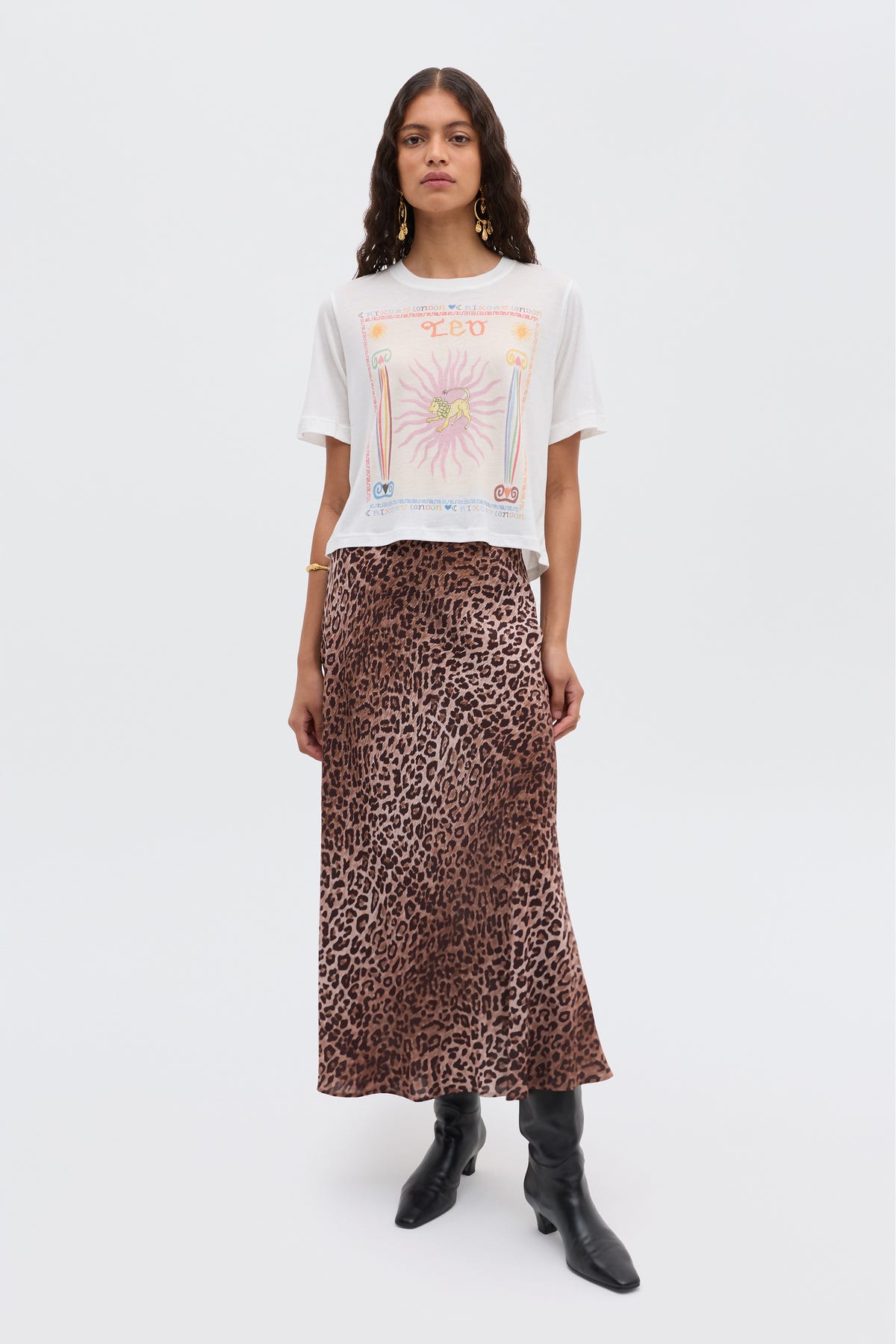 A woman with long wavy hair and a neutral expression stands against a plain background, wearing a white graphic tee, the Ria - Leo skirt with cropped hem, black knee-high boots, and gold earrings and bracelet.