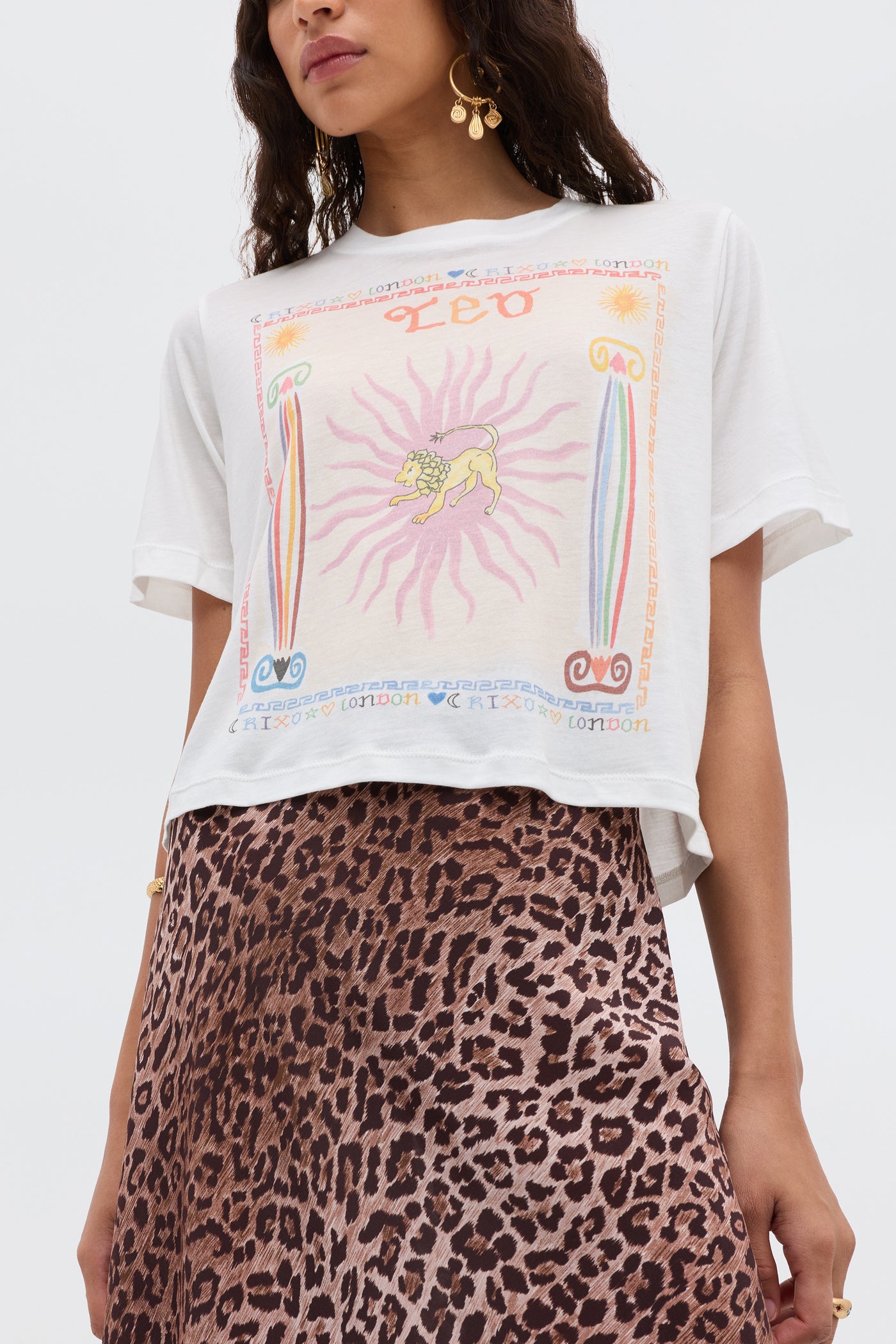 Woman wears the Ria - Leo T-shirt, featuring a colorful Leo illustration, paired with a brown leopard print RIXO skirt and gold accessories, standing against a plain background.