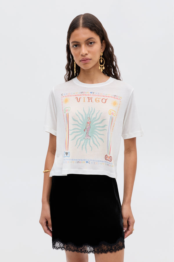 A woman with long wavy hair wears the Ria - Virgo white t-shirt with a colorful Virgo design and a black velvet skirt with lace trim, standing against a plain light background.