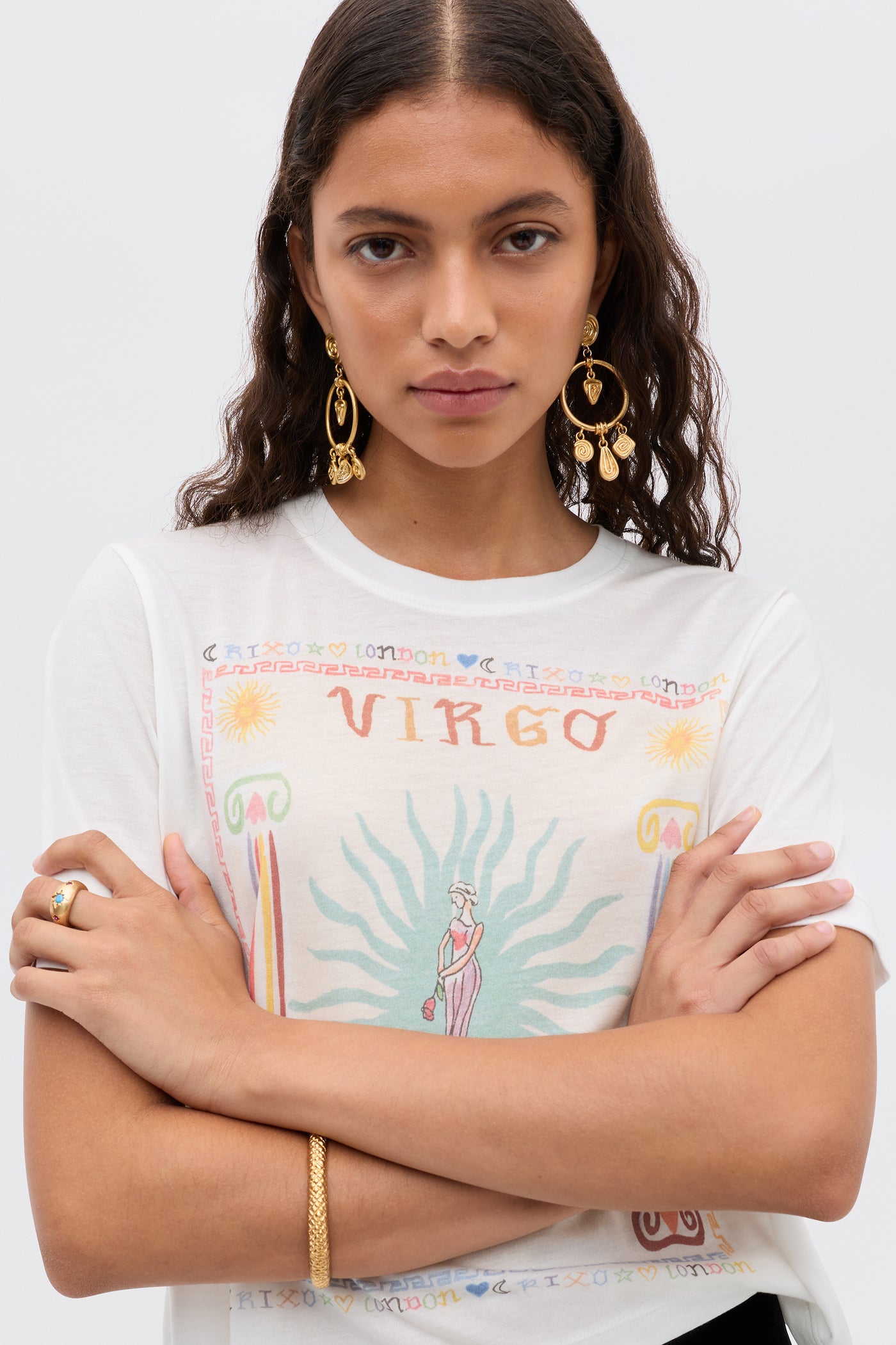 A woman with wavy brown hair wears gold earrings and a white Ria - Virgo T-shirt featuring a colorful Virgo illustration and cropped hem, standing arms crossed against a plain background—perfect to pair with RIXO skirts.