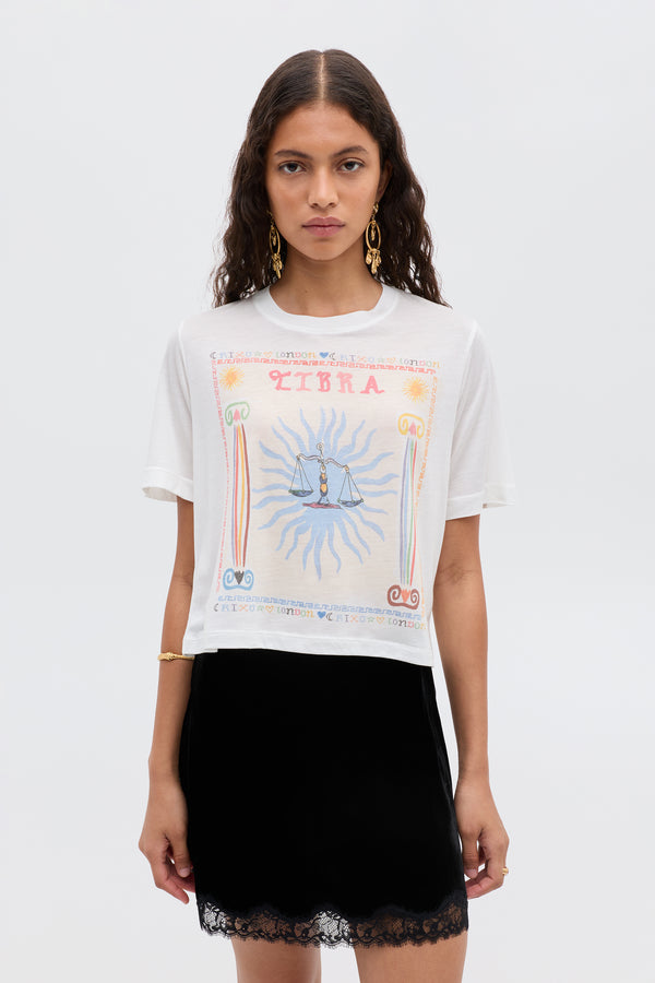 A woman with wavy brown hair wears the Ria - Libra white T-shirt with a colorful Libra illustration and a short black skirt, standing against a plain light background.