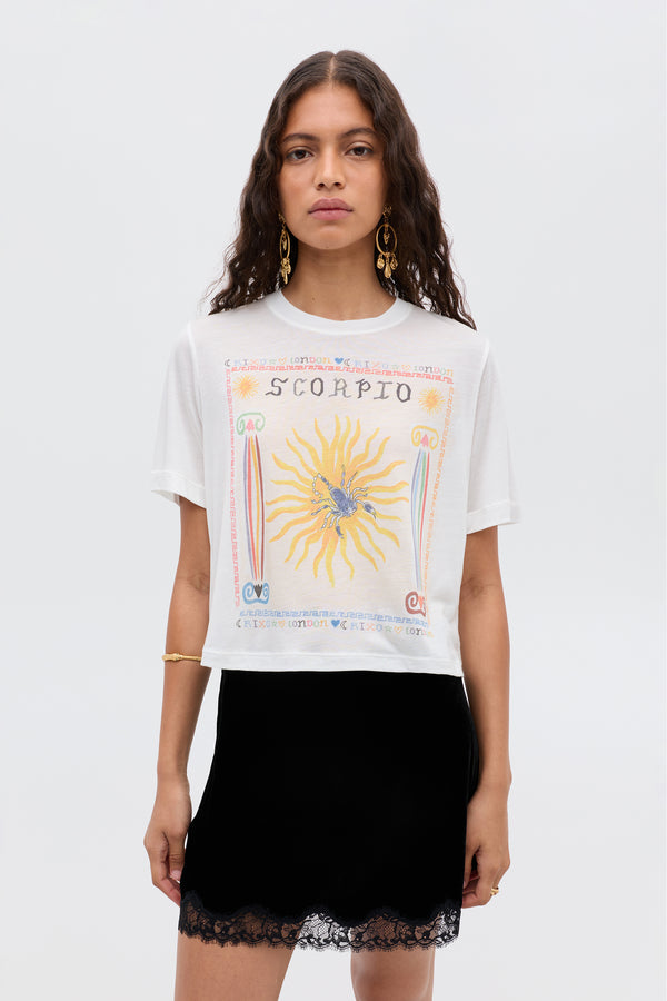 A woman with wavy brown hair wears the Ria - Scorpio white t-shirt with a sun and Scorpio graphic, paired with a lace-trimmed, cropped-hem RIXO skirt. She stands facing forward against a plain light background.