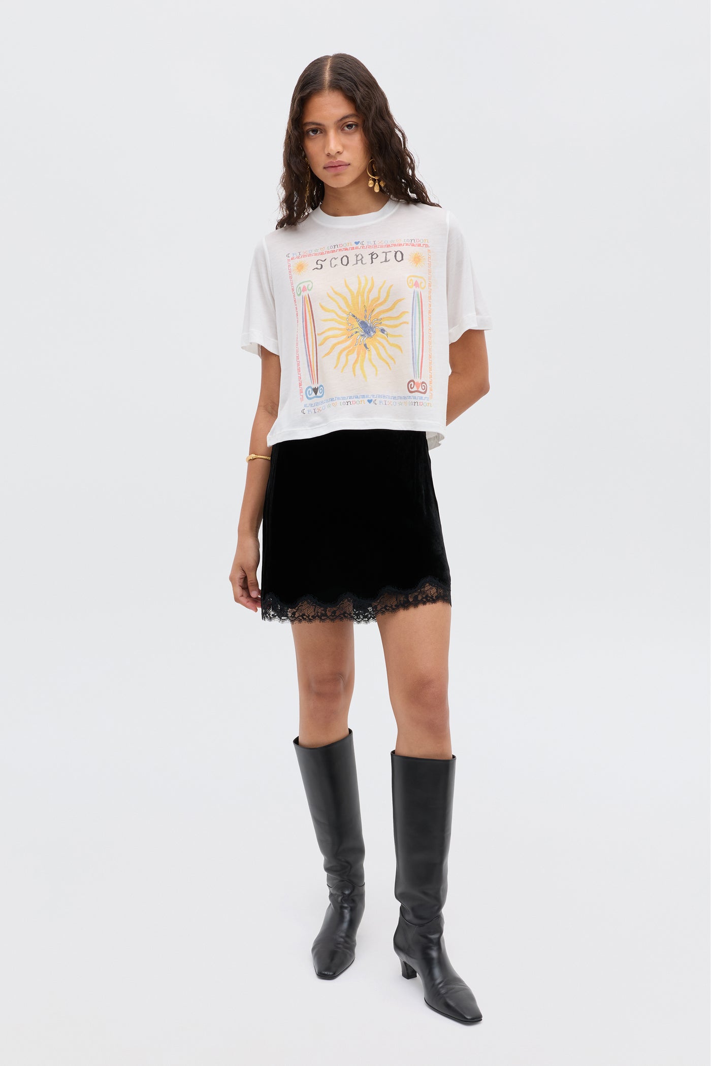 A woman with long wavy hair wears the Ria - Scorpio white graphic T-shirt with a bold Scorpio illustration, a lace-trimmed, cropped-hem RIXO skirt, and knee-high black boots, standing against a plain light background.