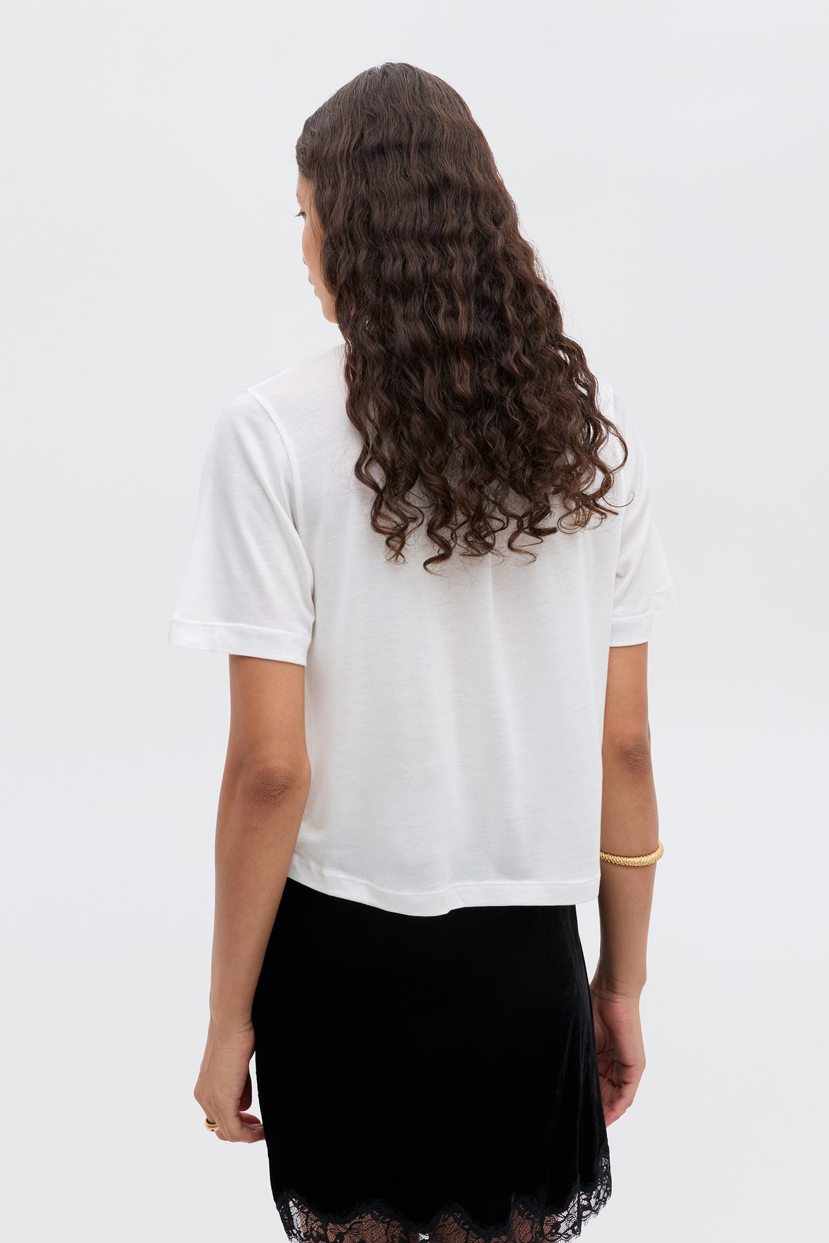 A person with long, curly brown hair is shown from the back, wearing a white short-sleeve shirt and the black RIXO "Ria - Scorpio" skirt with lace trim. A gold bracelet adorns their right wrist against a plain light background.