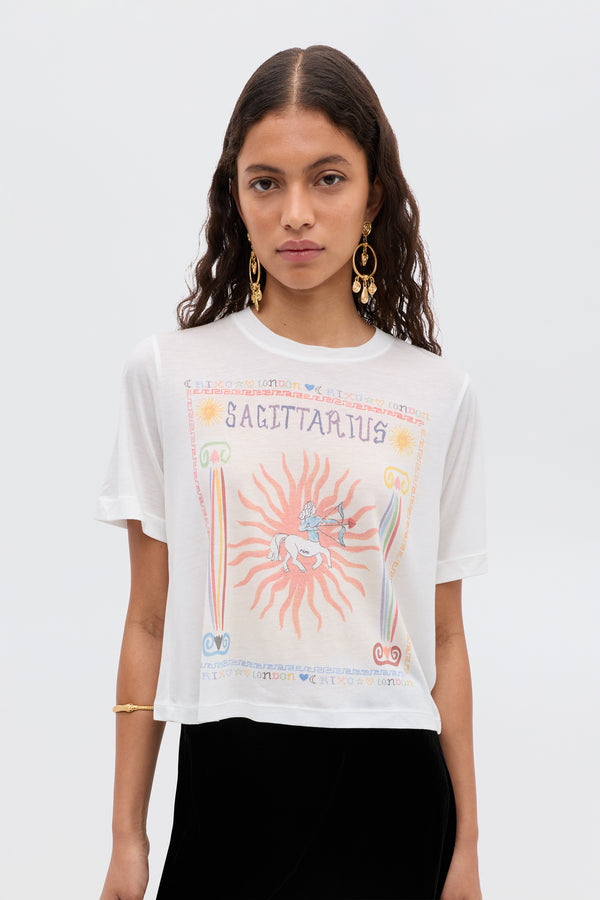 A woman in the Ria - Sagittarius white T-shirt, featuring a vivid centaur archer and sunburst graphic, styles its cropped hem with stylish RIXO skirts. She accessorizes with long wavy hair, gold earrings, and an arm bracelet.
