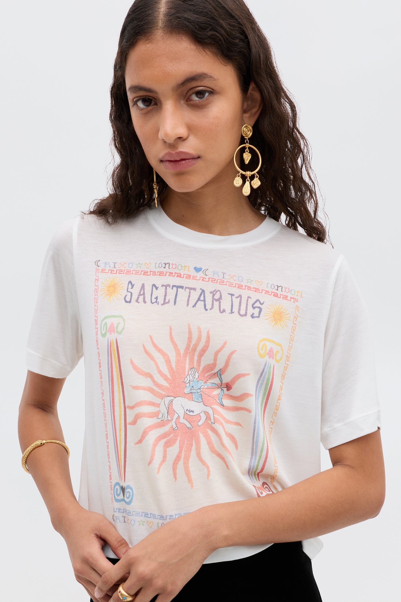 A woman with long wavy brown hair wears gold earrings and the Ria - Sagittarius tee, featuring a colorful centaur, sun, and borders. Its cropped hem pairs perfectly with RIXO skirts as she looks to the side with a neutral expression.