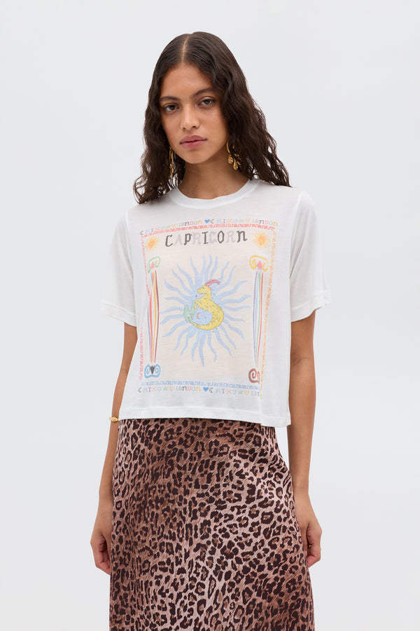 A woman with wavy brown hair wears the white Ria - Capricorn T-shirt, styled with a chic brown leopard print skirt, standing against a plain white background.