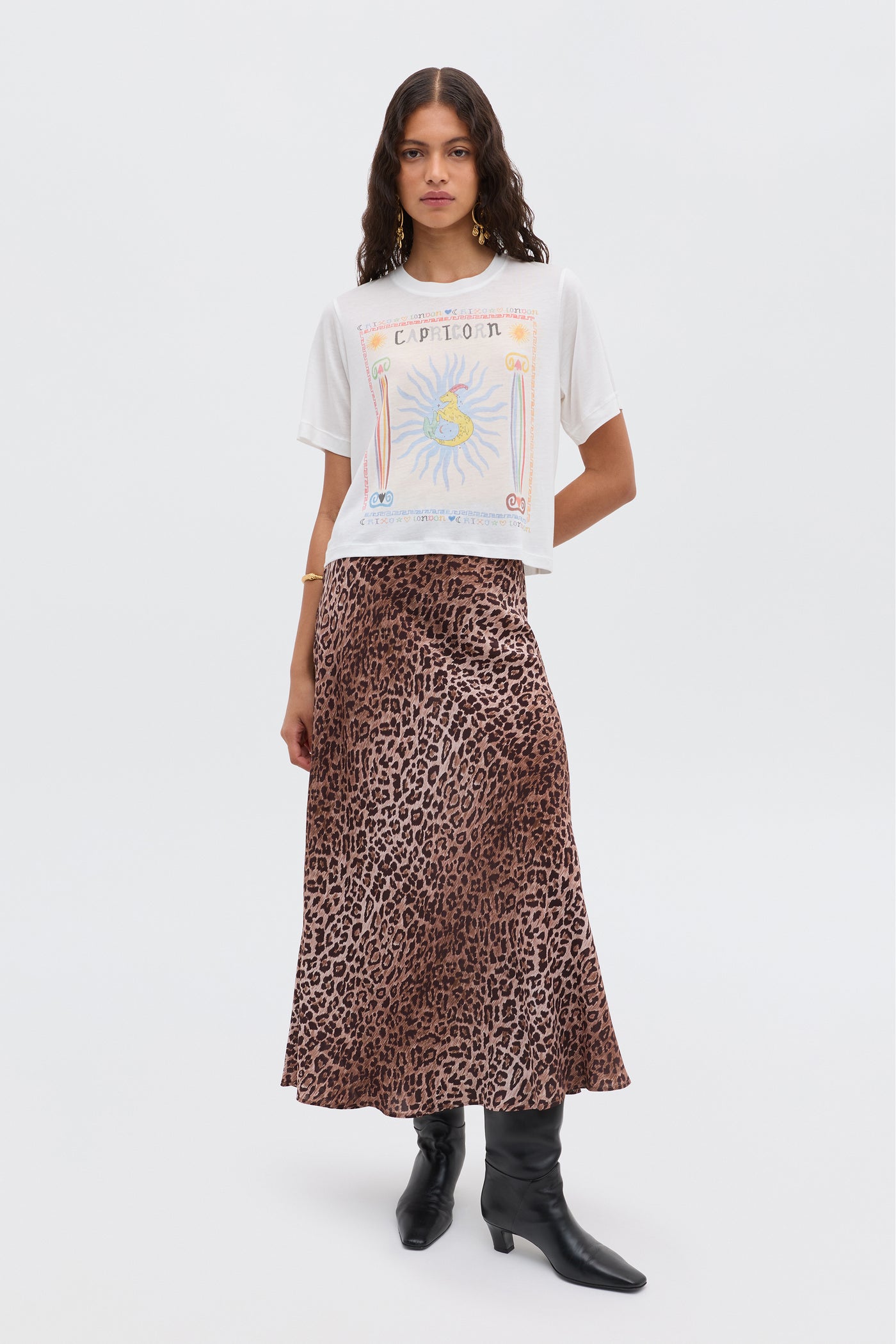 A woman with wavy brown hair and minimal accessories stands against a white background wearing the Ria - Capricorn white T-shirt, a brown leopard print skirt, and black knee-high boots.