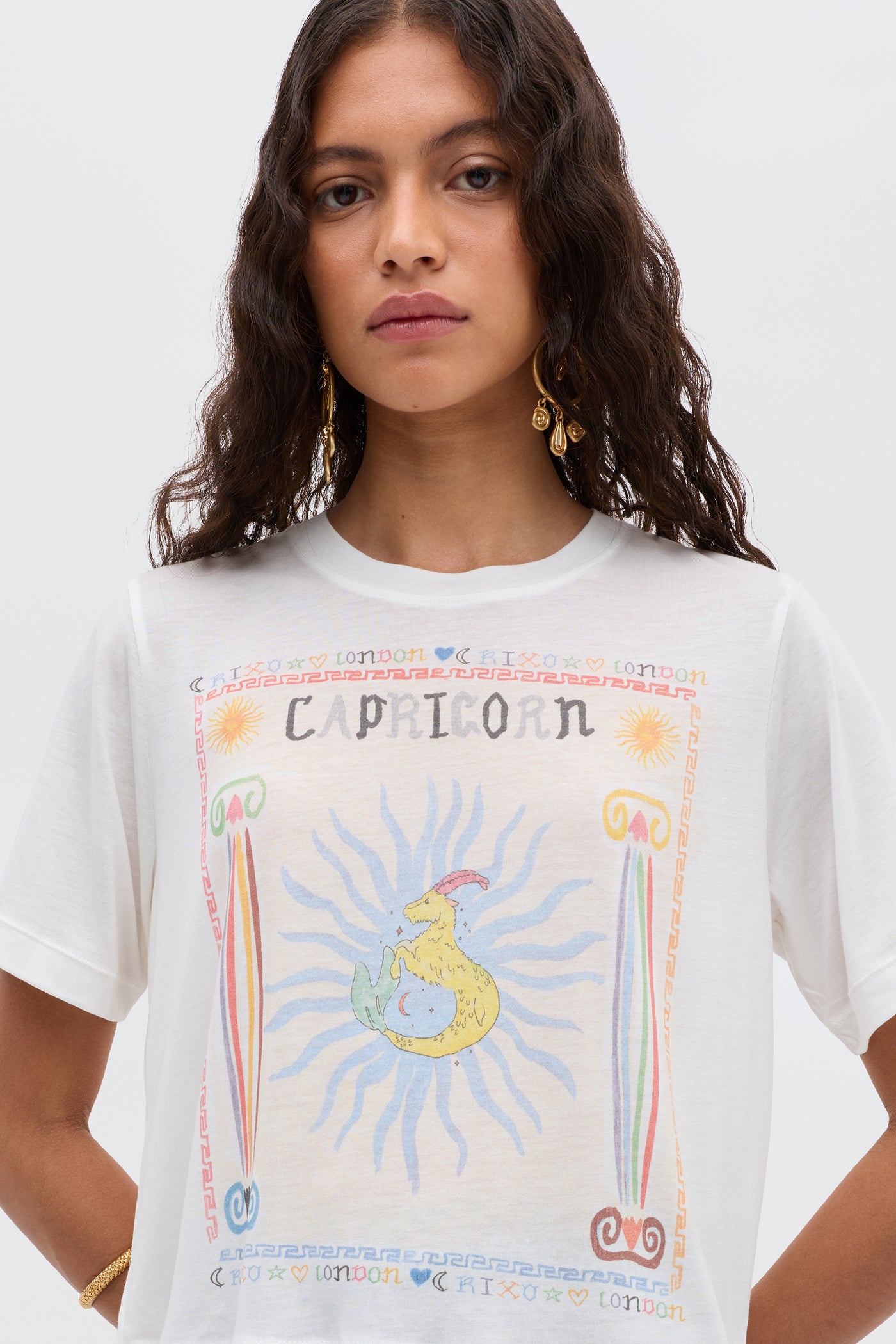 Woman with wavy brown hair wears gold earrings and the Ria - Capricorn T-shirt, featuring a vibrant Capricorn illustration, symbols, and "CAPRICORN" and "LONDON" text—styled chicly with RIXO skirts for an astrological look.