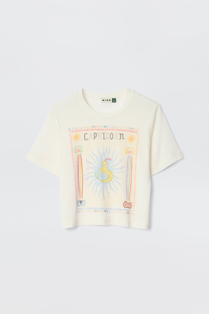 The Ria - Capricorn white short-sleeve T-shirt showcases a bold Capricorn graphic with a sun, astrological symbols, and decorative borders on the front. Ideal with RIXO skirts, it's photographed on a plain light background.