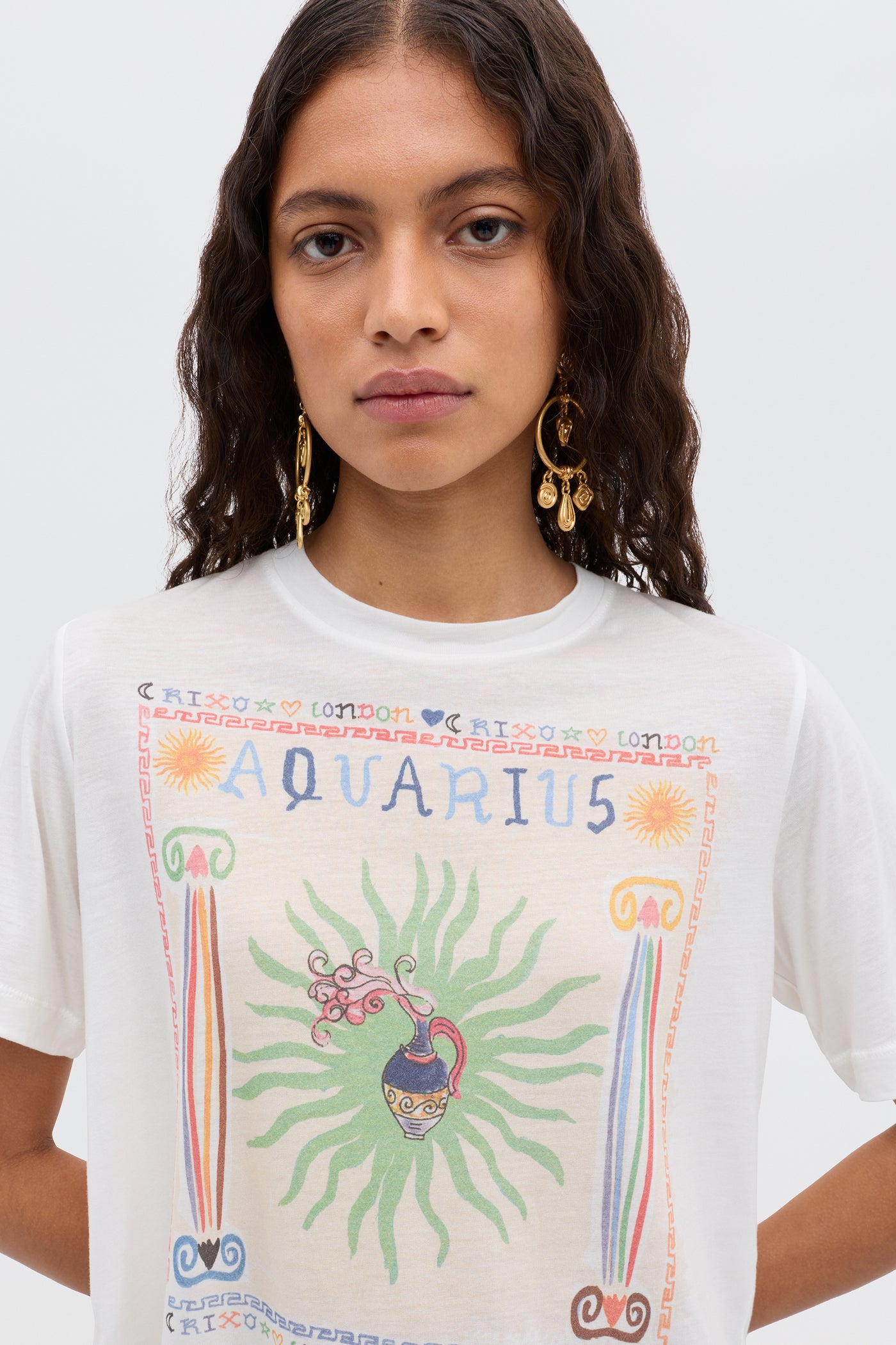 A woman with wavy brown hair wears gold dangle earrings and the Ria - Aquarius white T-shirt, featuring sun motifs, green rays, Greek-style columns, and a cropped hem—perfect with chic RIXO skirts.