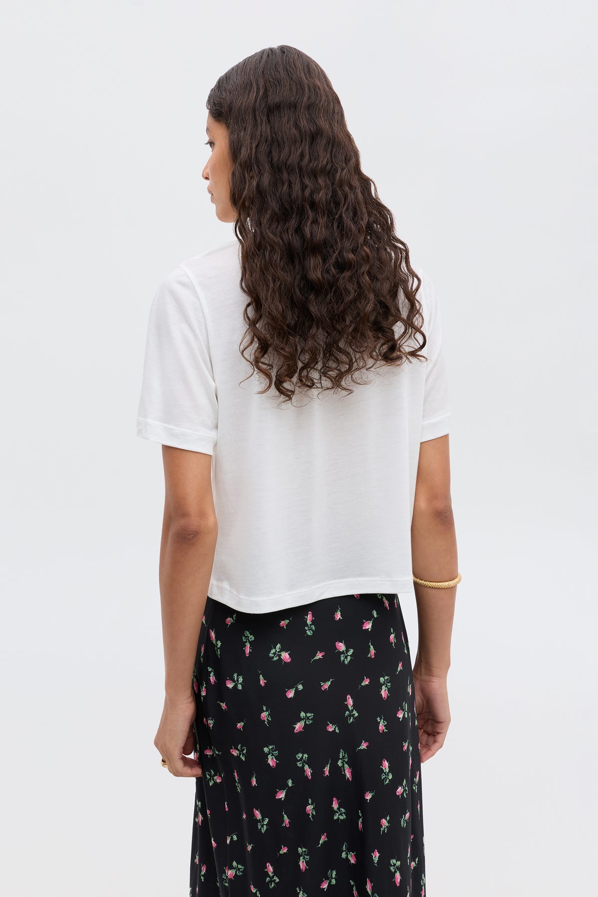 A person with long curly hair, in a white short-sleeve shirt and the black RIXO "Ria - Aquarius" skirt with a cropped hem, is standing with their back to the camera against a plain light background.