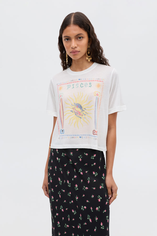 A woman with wavy dark hair wears the Ria - Pisces white cropped hem T-shirt with a colorful Pisces illustration and a black floral skirt, standing against a plain white background.