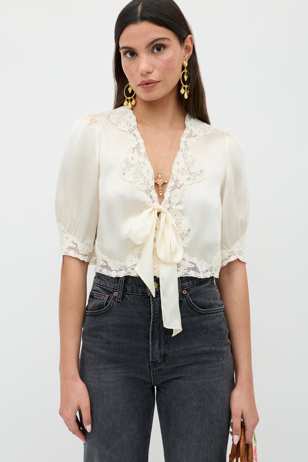 A woman in the elegant Amara - Champagne top—a cream satin blouse with lace trim and a bow—pairs it with high-waisted dark jeans. She has long dark hair, gold earrings, and a neutral look, standing against a plain background.