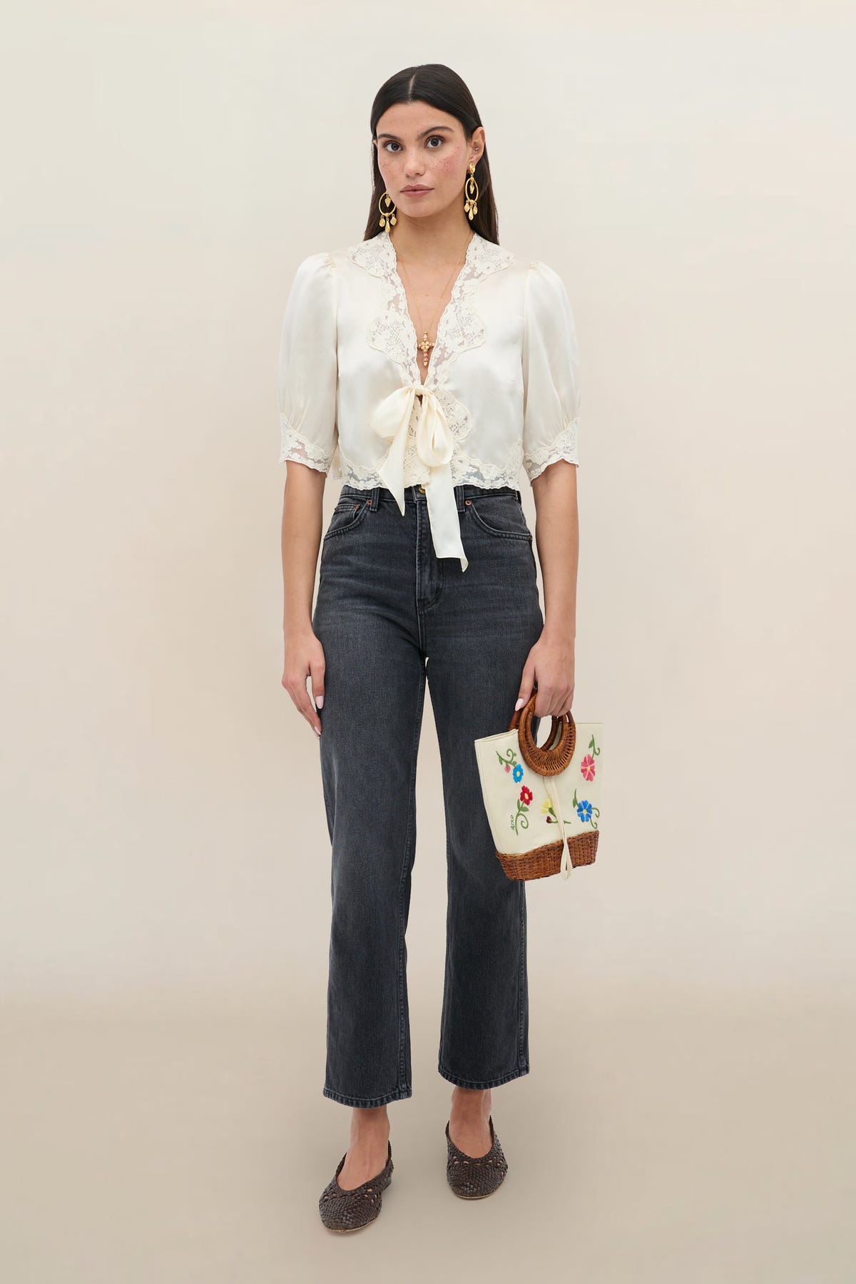 A woman stands against a plain background wearing the Amara - Champagne silk-satin top with lace trim and bow, paired with high-waisted dark jeans, brown shoes, gold earrings, and a woven handbag with embroidered flowers.