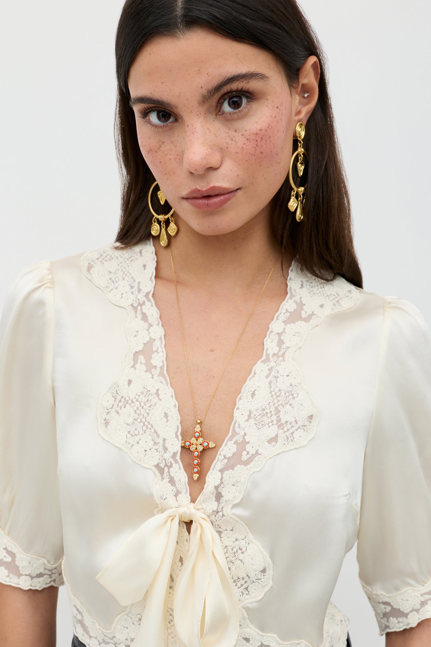 A woman with long dark hair and freckles wears the Amara - Champagne top, featuring lace trim and a bow, along with gold drop earrings and a gold cross necklace. She faces forward against a plain light background.