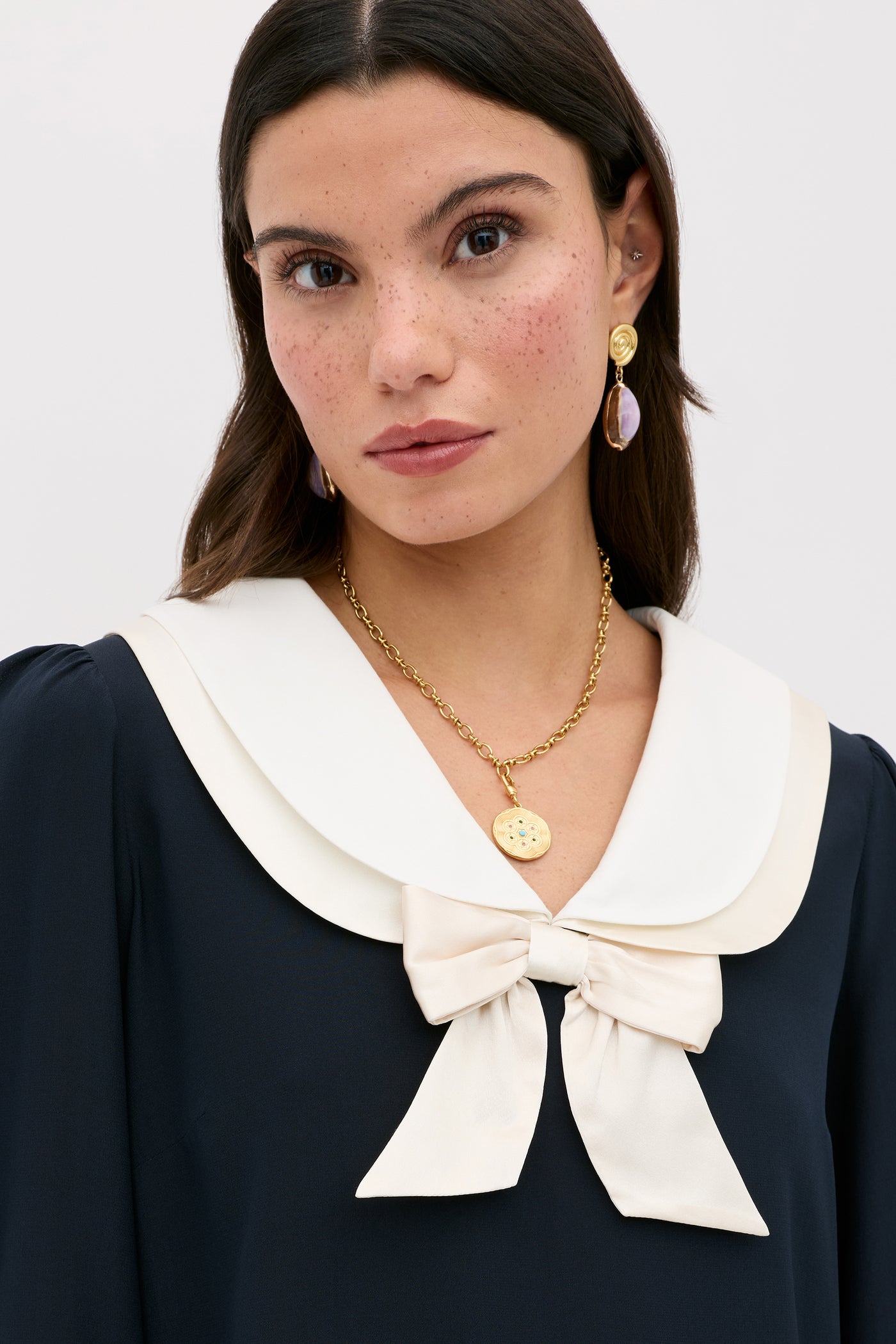 A woman with dark hair wears the Eimer - Navy, a silk top with a sailor-style collar and large bow. She accessorizes with gold earrings and a round pendant necklace, posing neutrally against a plain light gray background.