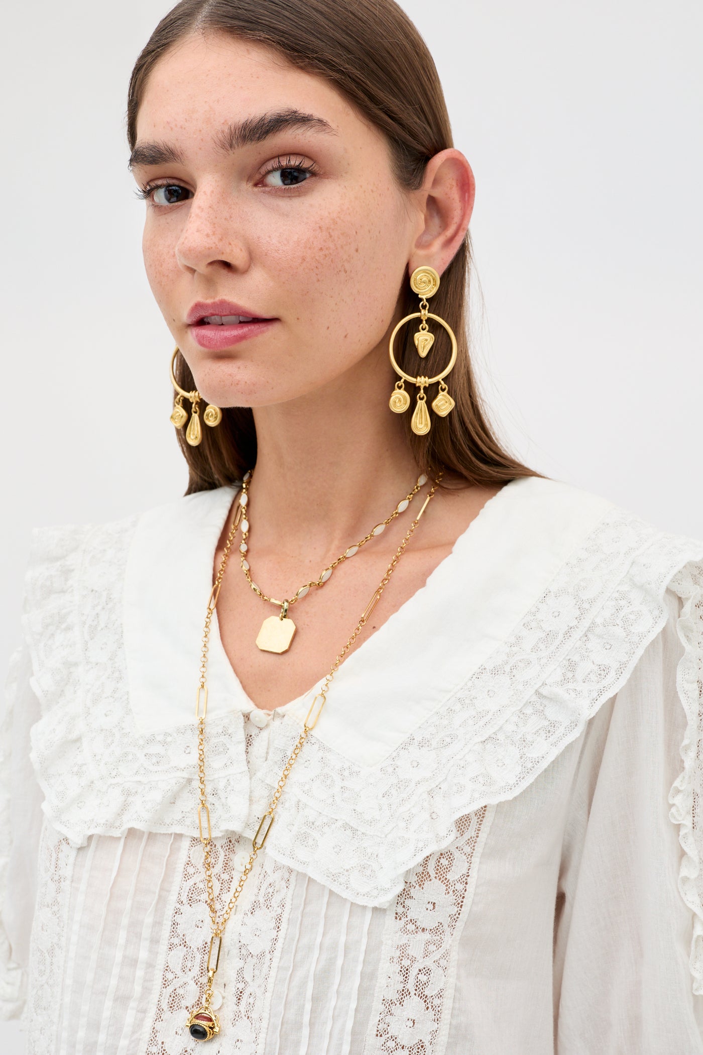 A woman with long brown hair wears the Ellery - White vintage-inspired cotton voile blouse with a ruffled collar. She accessorizes with gold hoop earrings, layered gold necklaces, and has a light freckled complexion and neutral expression.