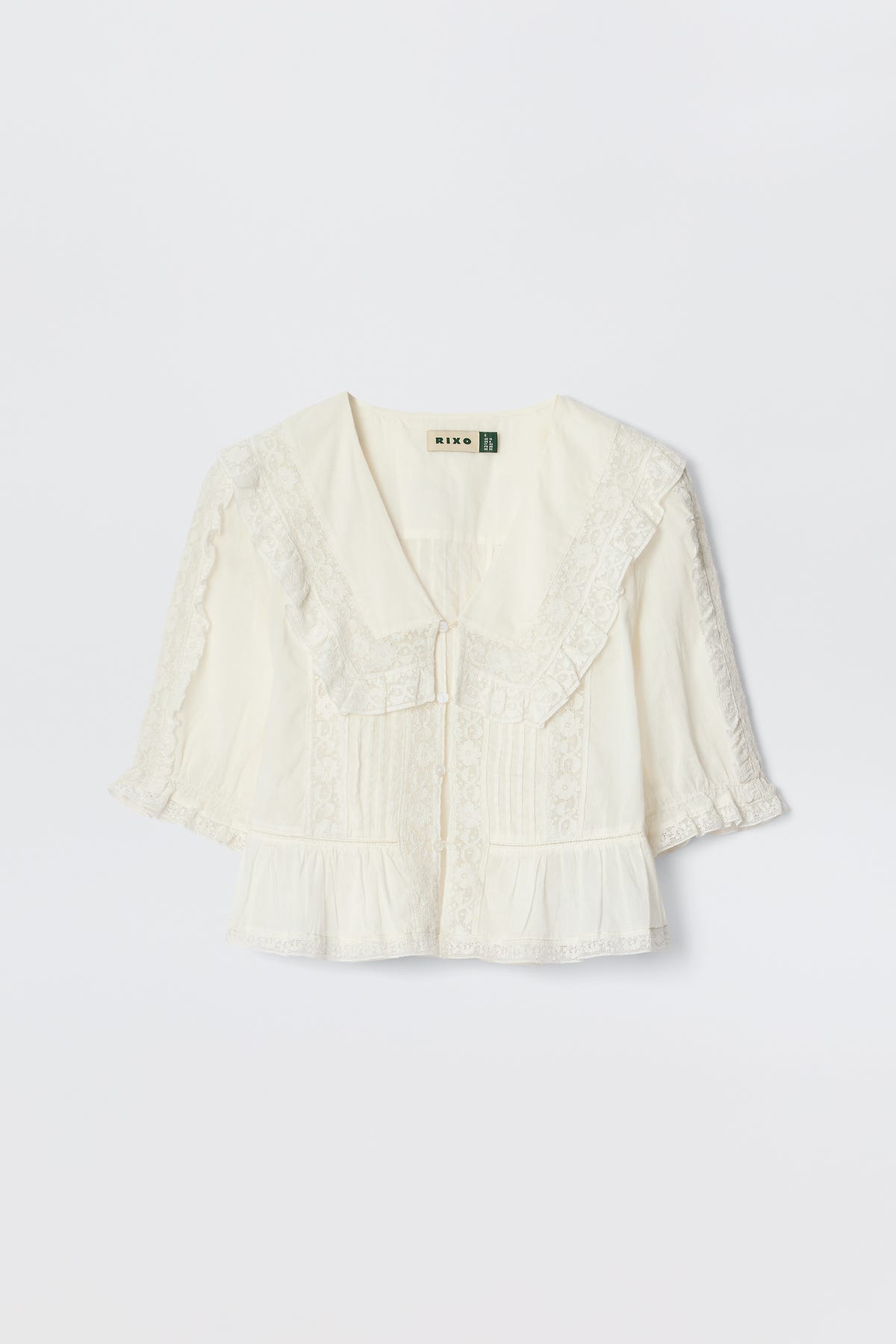 The Ellery - White is a cream-colored, vintage-inspired blouse made from lightweight cotton voile with a wide ruffled lace collar, short puff sleeves, front pleats, and delicate lace trim, shown on a plain white background.