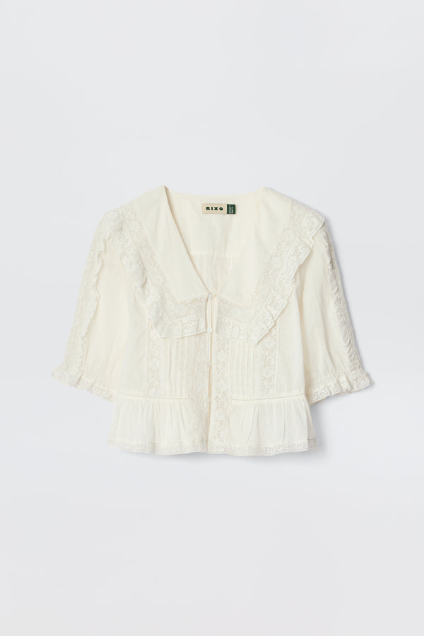 The Ellery - White is a cream-colored, vintage-inspired blouse made from lightweight cotton voile with a wide ruffled lace collar, short puff sleeves, front pleats, and delicate lace trim, shown on a plain white background.