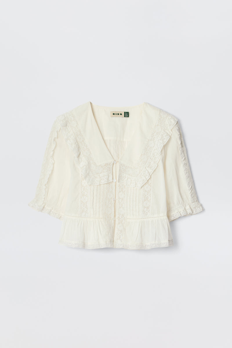 The Ellery - White is a cream-colored, vintage-inspired blouse made from lightweight cotton voile with a wide ruffled lace collar, short puff sleeves, front pleats, and delicate lace trim, shown on a plain white background.
