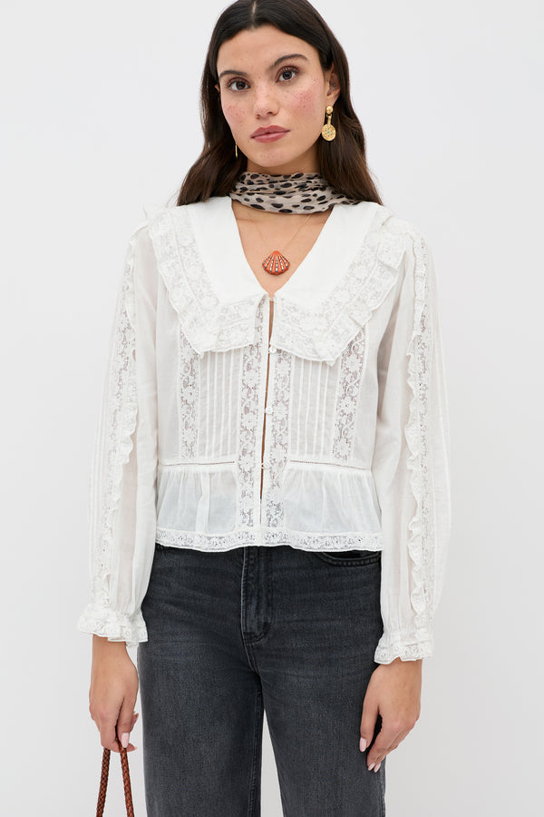 A woman stands against a white background wearing the Maribelle - Ivory vintage-inspired top with a large collar, paired with black jeans, a leopard print scarf, gold earrings, and holding a brown bag.