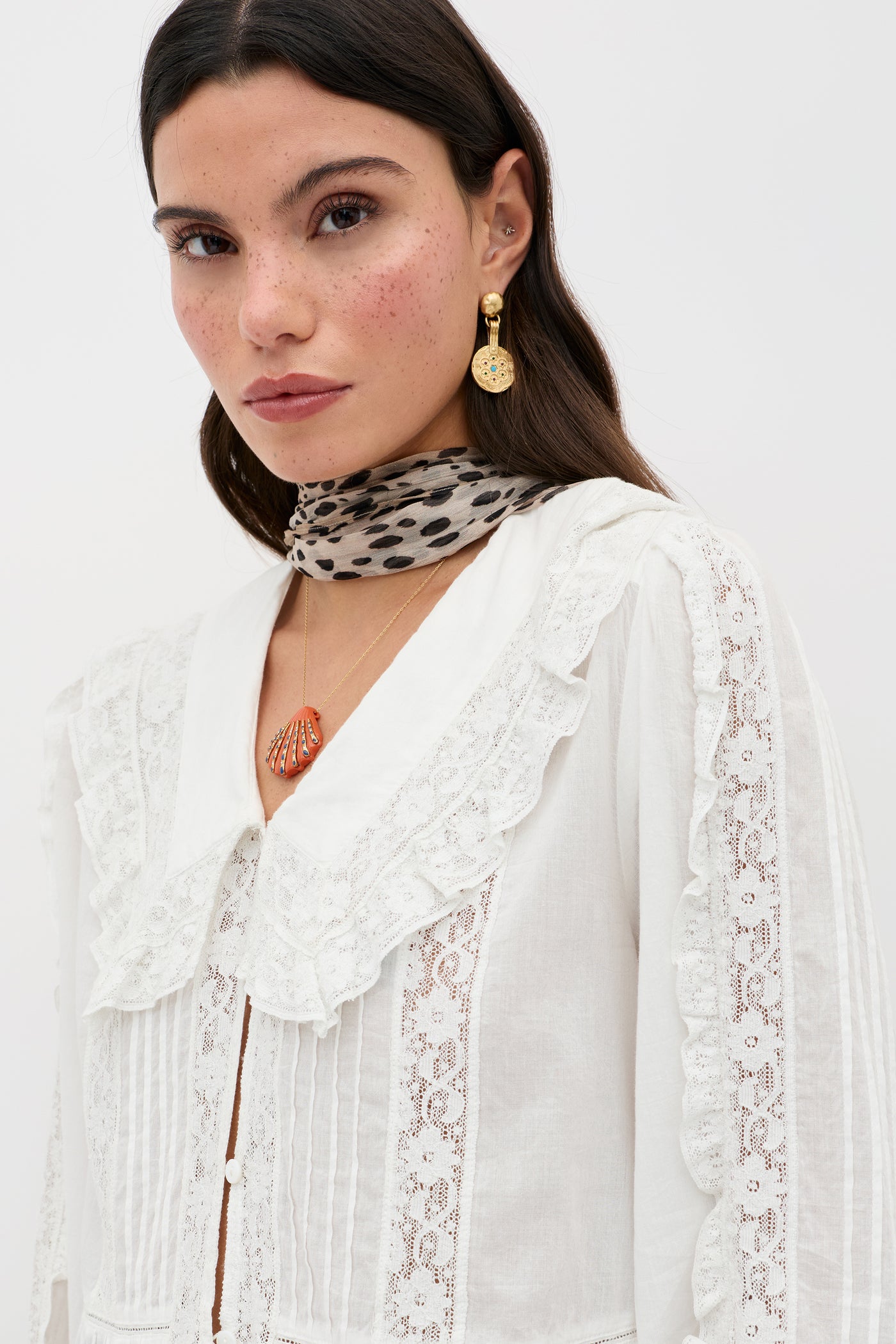 A woman with long dark hair and freckles wears the Maribelle - Ivory, a vintage-inspired white lace blouse with a large ruffled collar, styled with a leopard print scarf, gold earrings, and a gold pendant. She has a neutral expression.