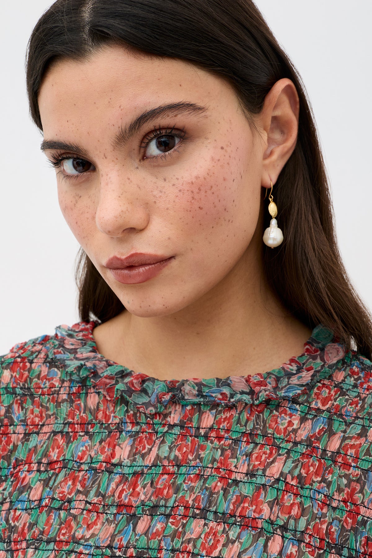 A woman with long brown hair, natural makeup, and freckles wears the Gardenia - Jeannie Bloom Charcoal floral top and a gold earring with a pearl drop, looking directly at the camera against a plain light background.