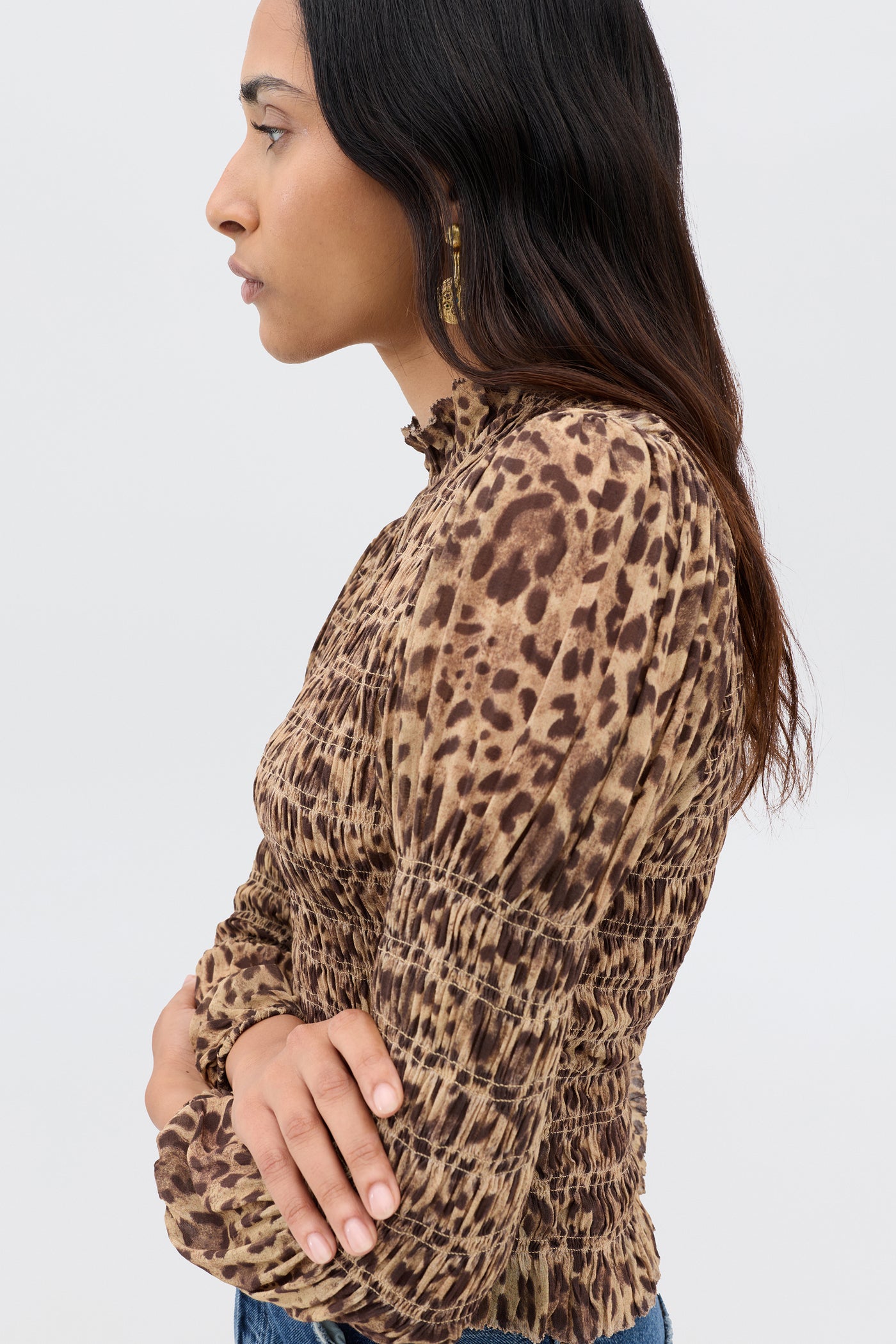 A woman with long dark hair stands in profile, wearing the Gardenia - Mila Leopard Brown top with long sleeves and ruched details. Her arms are crossed as she looks forward against a plain light background.