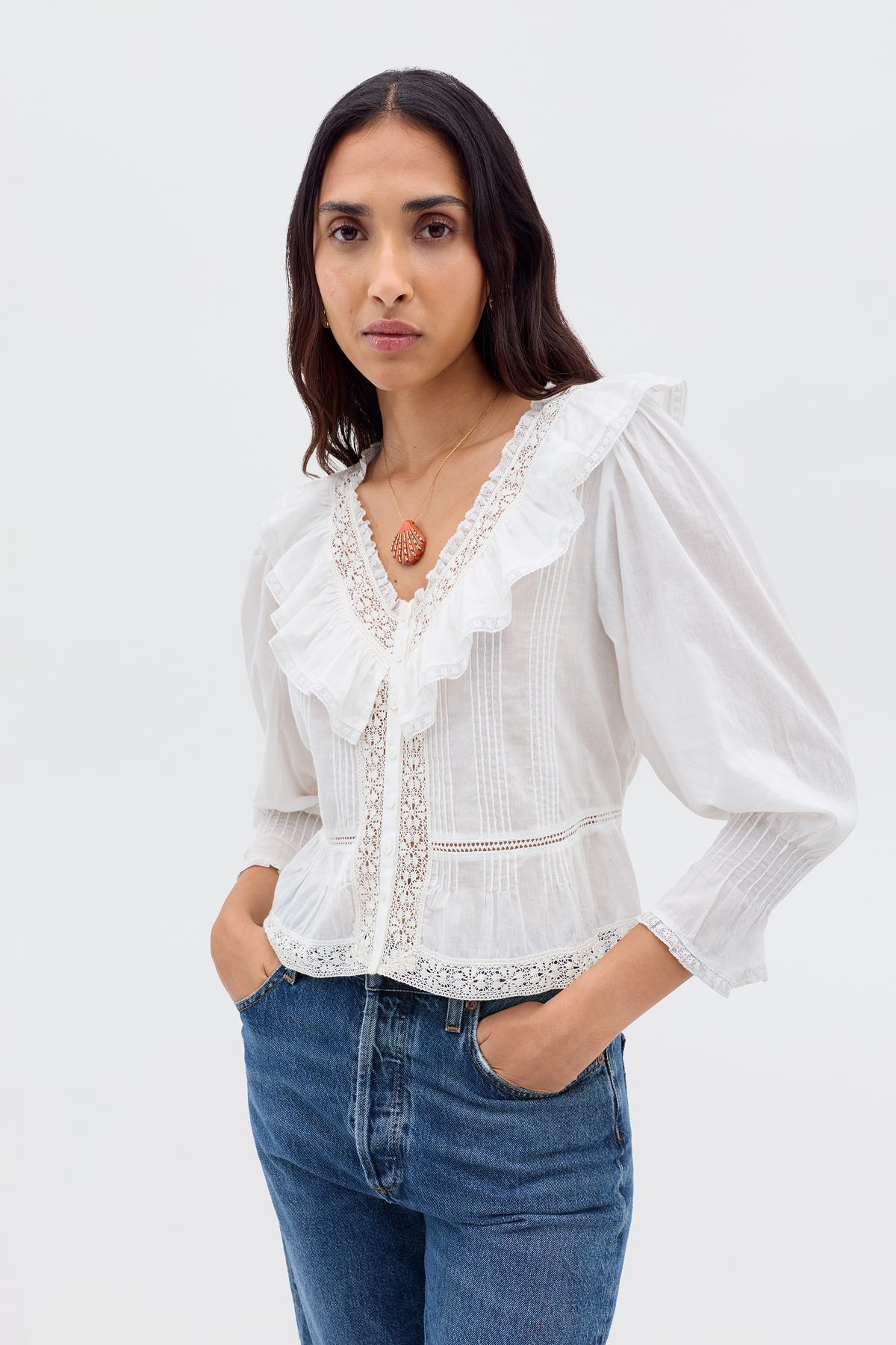 A woman with long brown hair wears the Solene - Ivory blouse—vintage-inspired with puffed sleeves, lace details, and a ruffled V-neckline—paired with blue jeans as she stands against a plain light background, hands in her pockets.