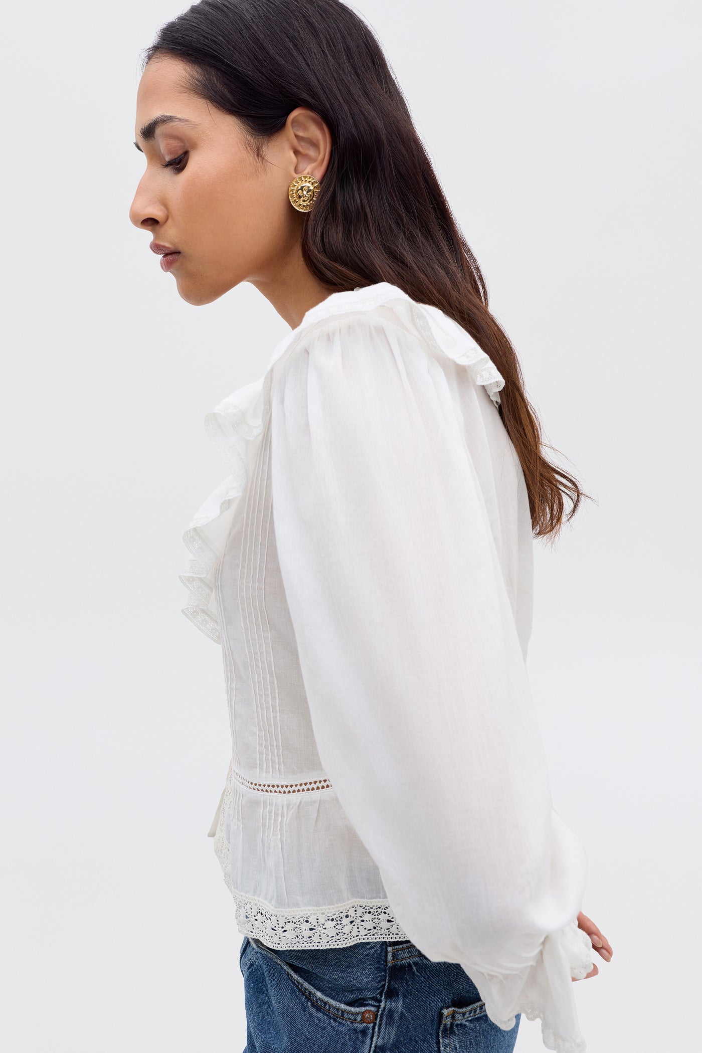 A woman with long dark hair, gold earrings, and blue jeans models the Solene - Ivory vintage-inspired blouse with puffed sleeves and a ruffled V-neckline, standing in profile against a plain light background.