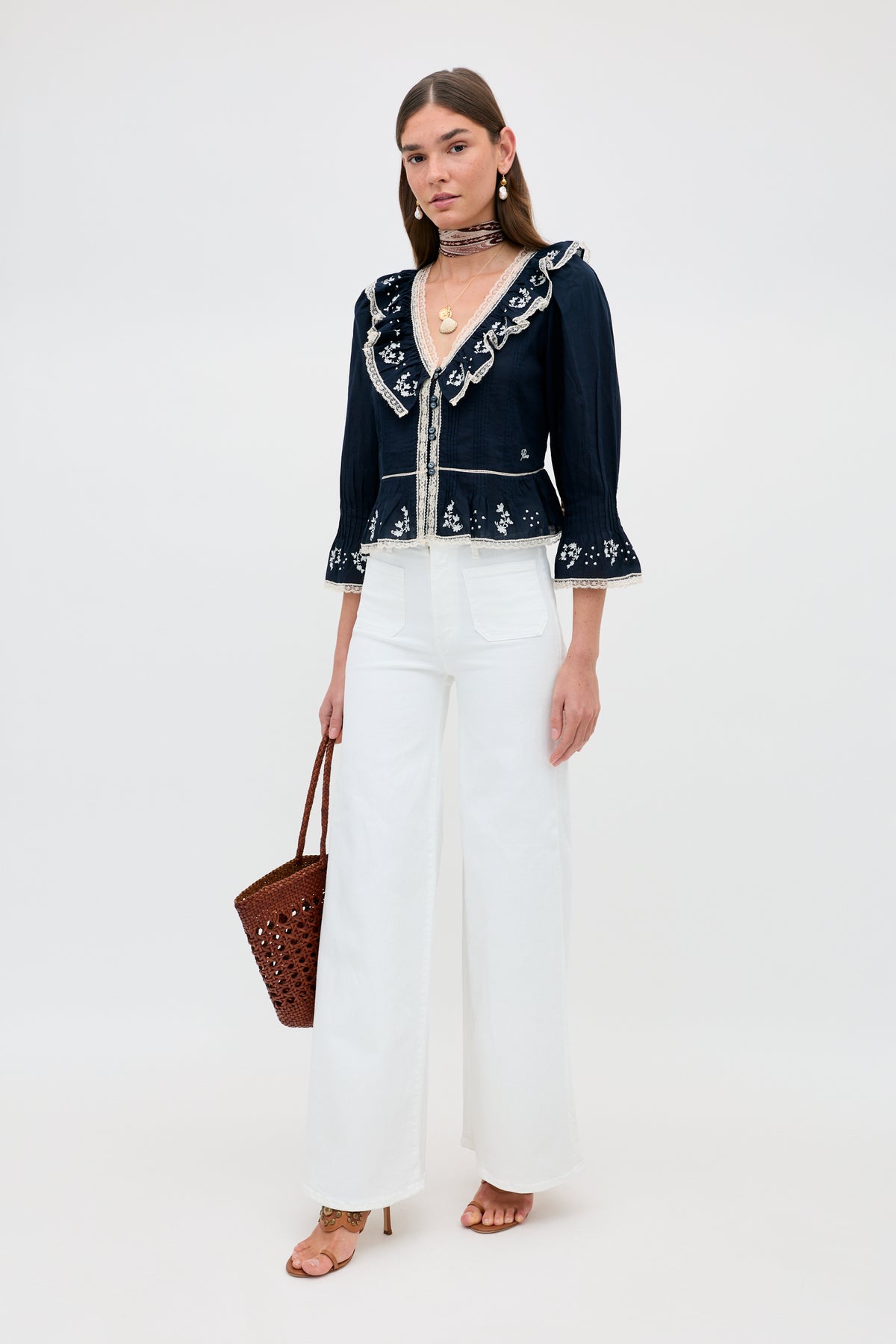 A woman with straight, long hair and gold jewelry wears the Solene - Flower Trail Embroidery Navy blouse with flared sleeves, white high-waisted wide-leg pants, tan heels, and carries a brown woven handbag for a timeless vintage look.
